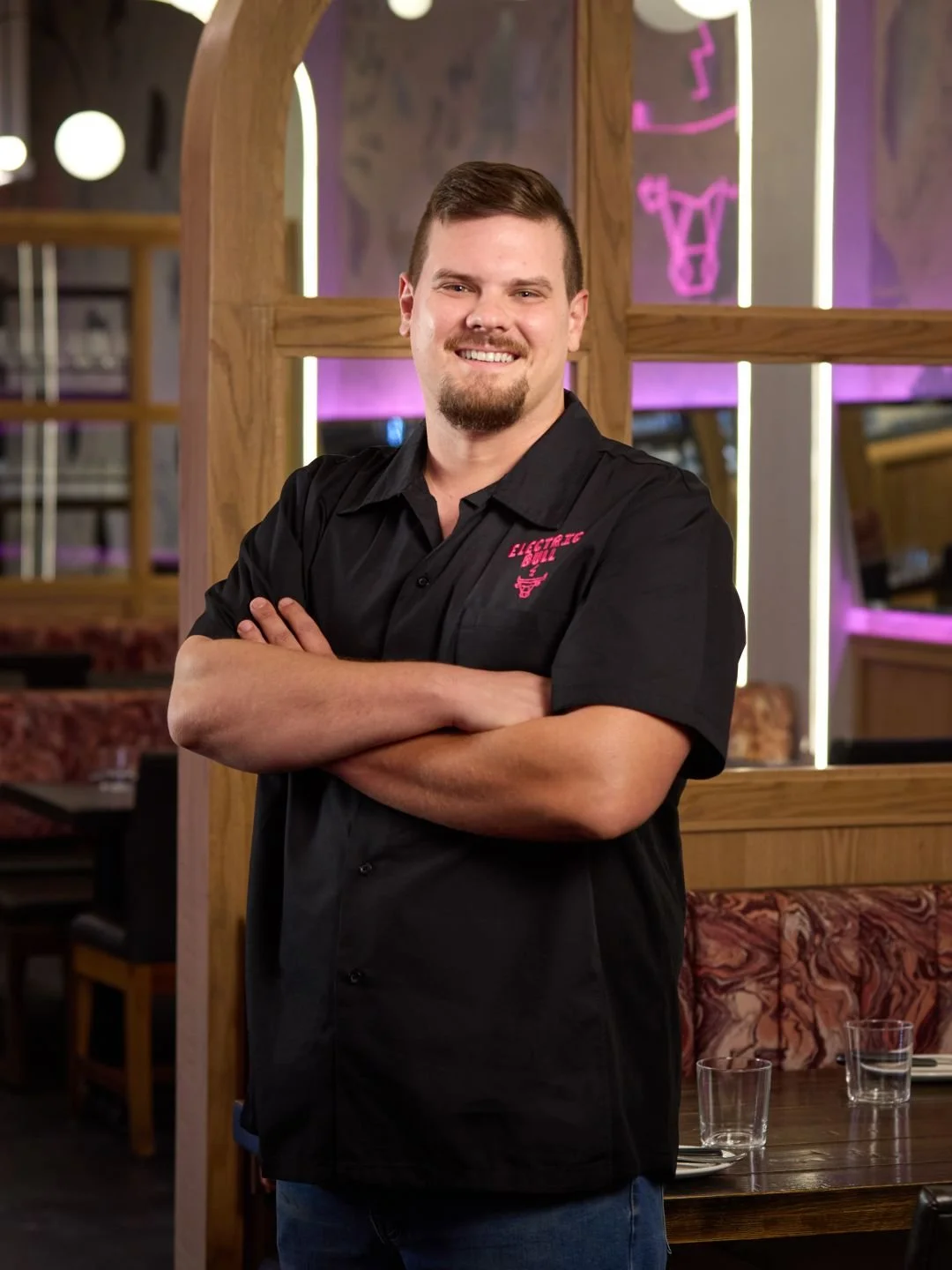 Head chef Adam Hoffa wearing a black shirt with pink embroidery that says 'Electric Bull" standing inside Electric Bull