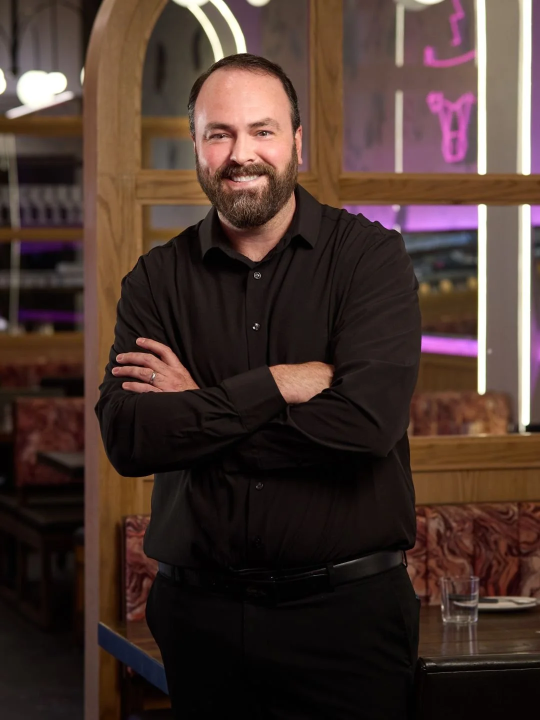 A man with a beard and short dark hair, wearing a black shirt, standing with arms crossed in a restaurant with modern decor and purple neon signs in the background.
