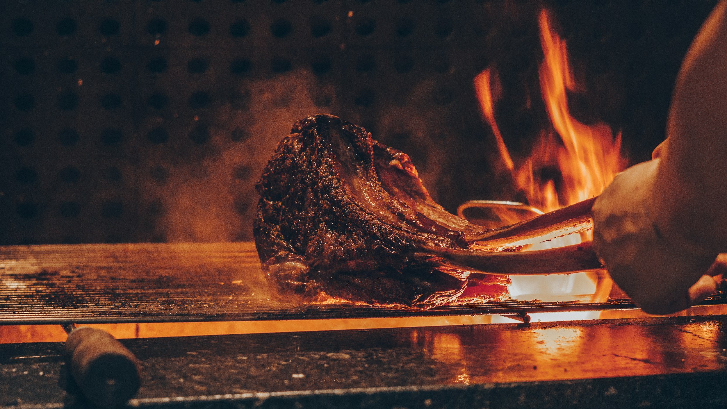A large piece of meat being cooked on a grill with flames, using tongs, in a dark kitchen or restaurant setting.