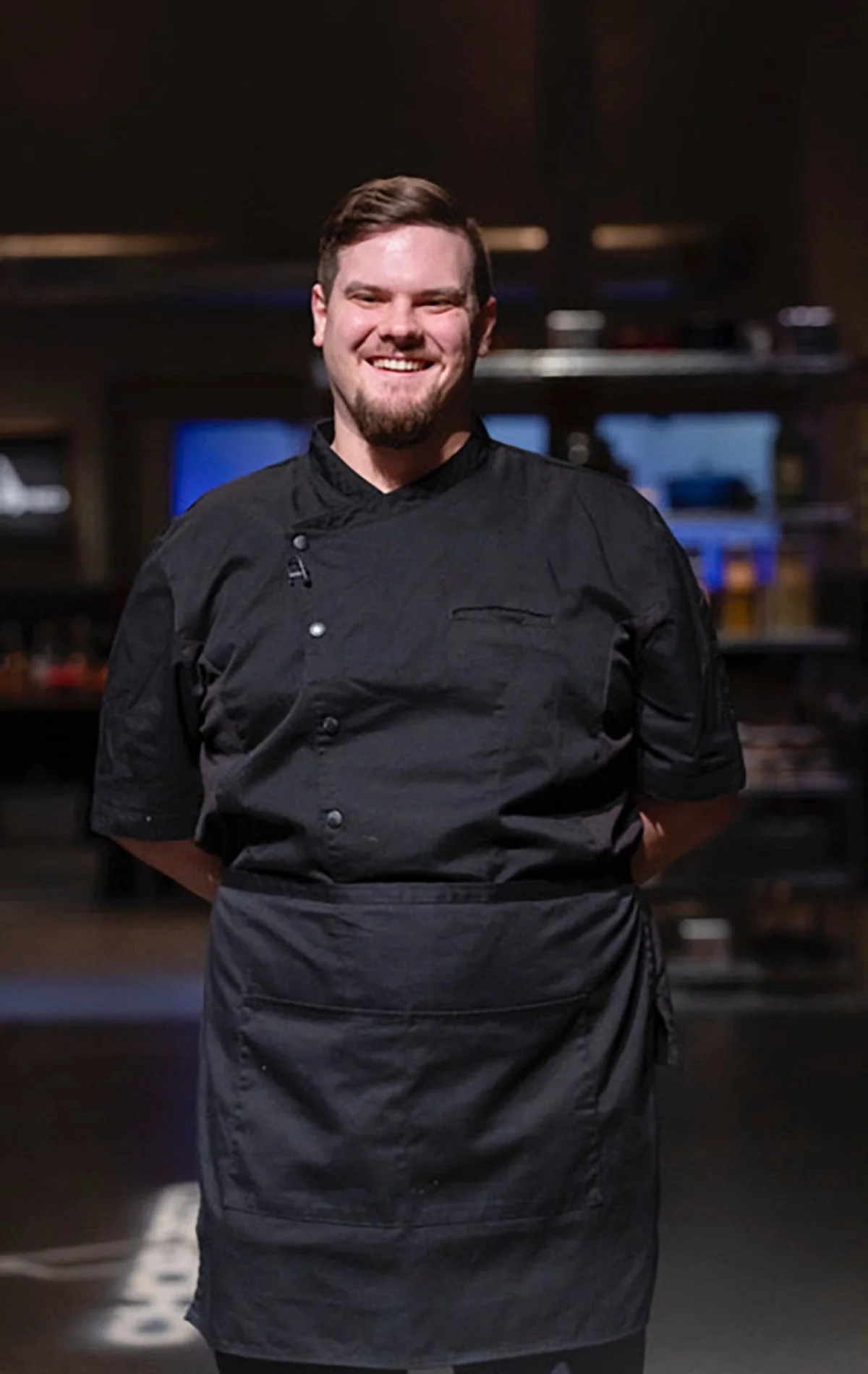 A smiling male chef wearing a black chef's coat and apron standing in a professional kitchen.