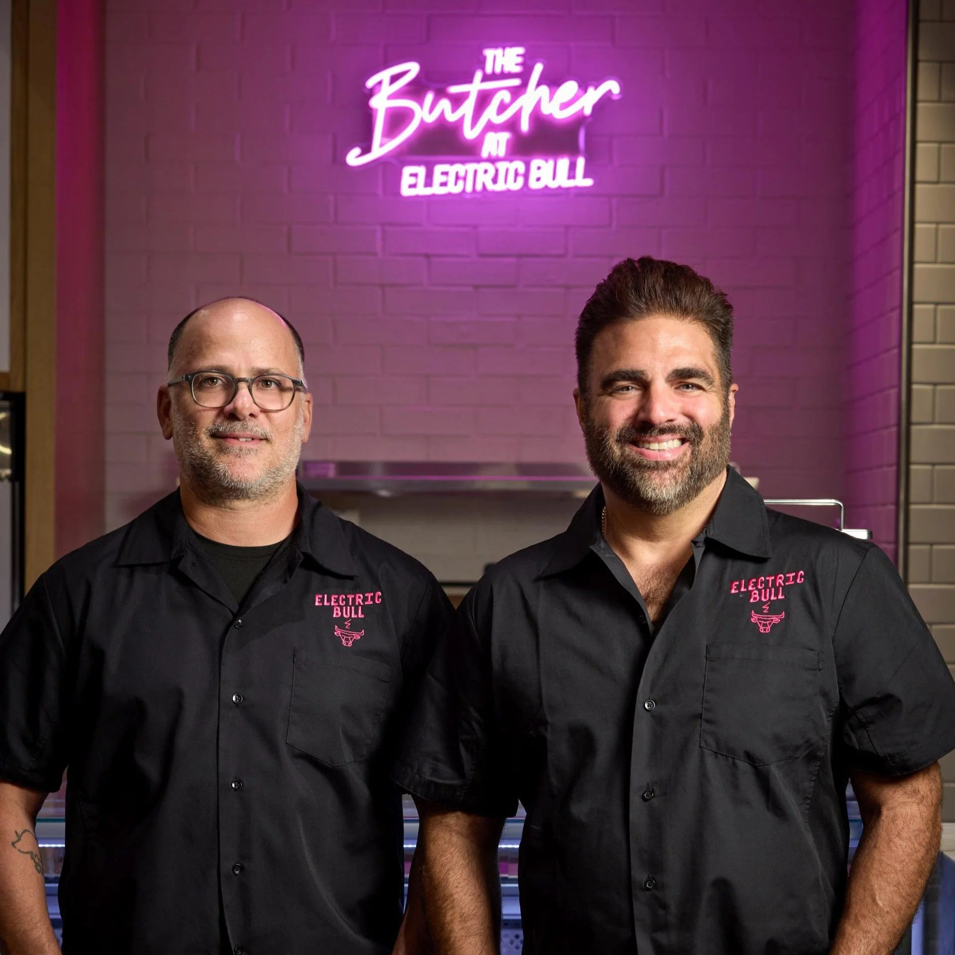 Chefs Victor Albisu and Justin Severino stand in front of a pink-purple neon sign that reads 'The Butcher at Electric Bull.'