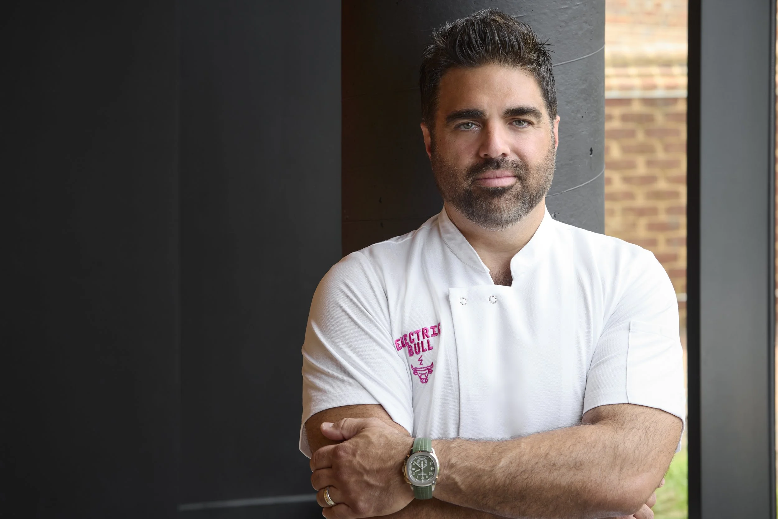 Chef Victor Albisu with dark hair and a beard, wearing a white chef's coat with 'Electric Bull' embroidered on the sleeve, standing indoors near a window with his arms crossed.