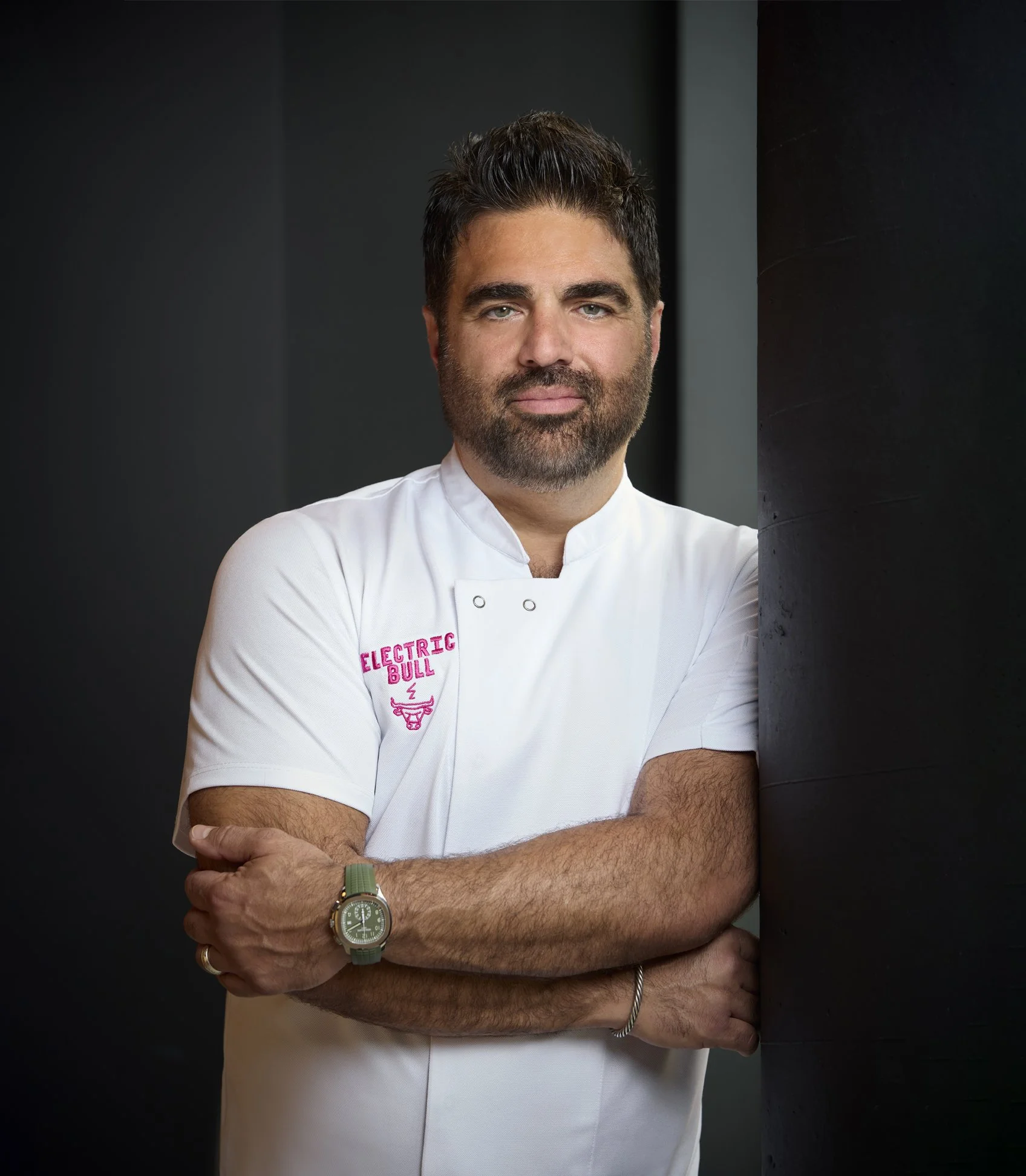 Chef Victor Albisu with dark hair and beard standing with arms crossed, wearing a white chef coat with pink embroidery that says 'Electric Bull', in front of a black wall.