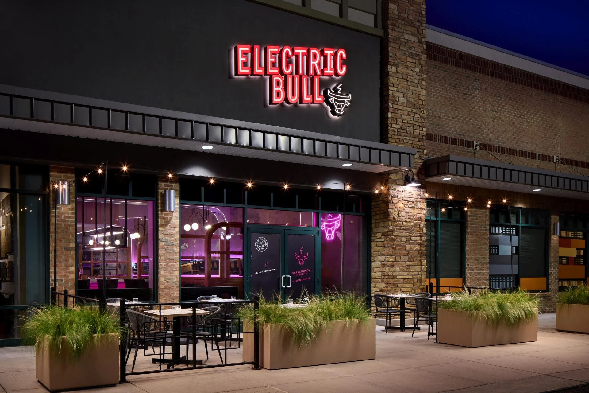 Exterior shot of Electric Bull featuring an outdoor patio, a large white and red sign with the letterhead and bull logo. You can see into the interior with led pink lighting.