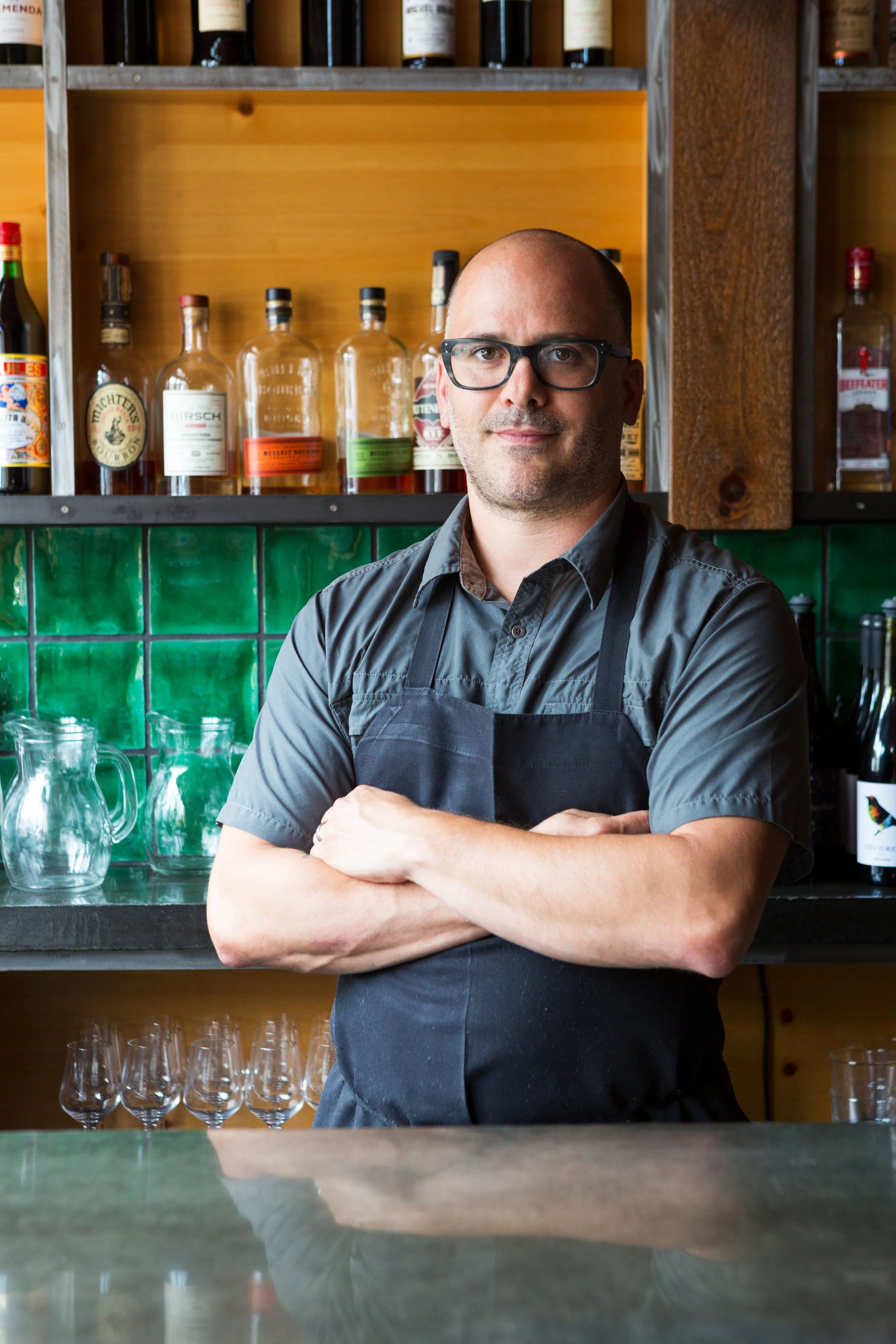 Chef Justin Severino wearing a gray shirt and black apron standing behind a bar with green tiled wall, bottles of alcohol, and glassware in the background.