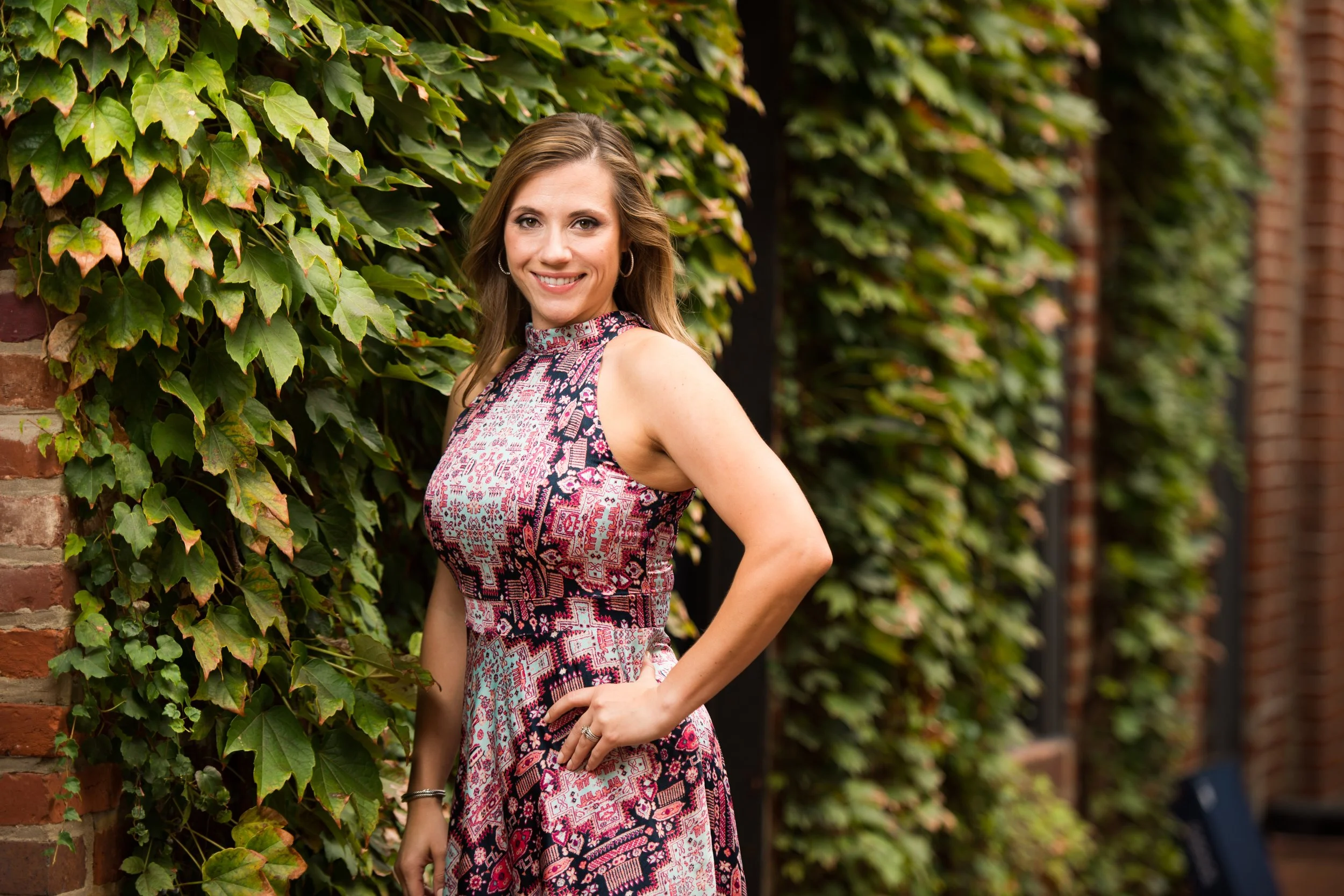 A woman with light brown hair in a patterned sleeveless dress standing beside a brick wall covered with green ivy leaves and smiling at the camera.