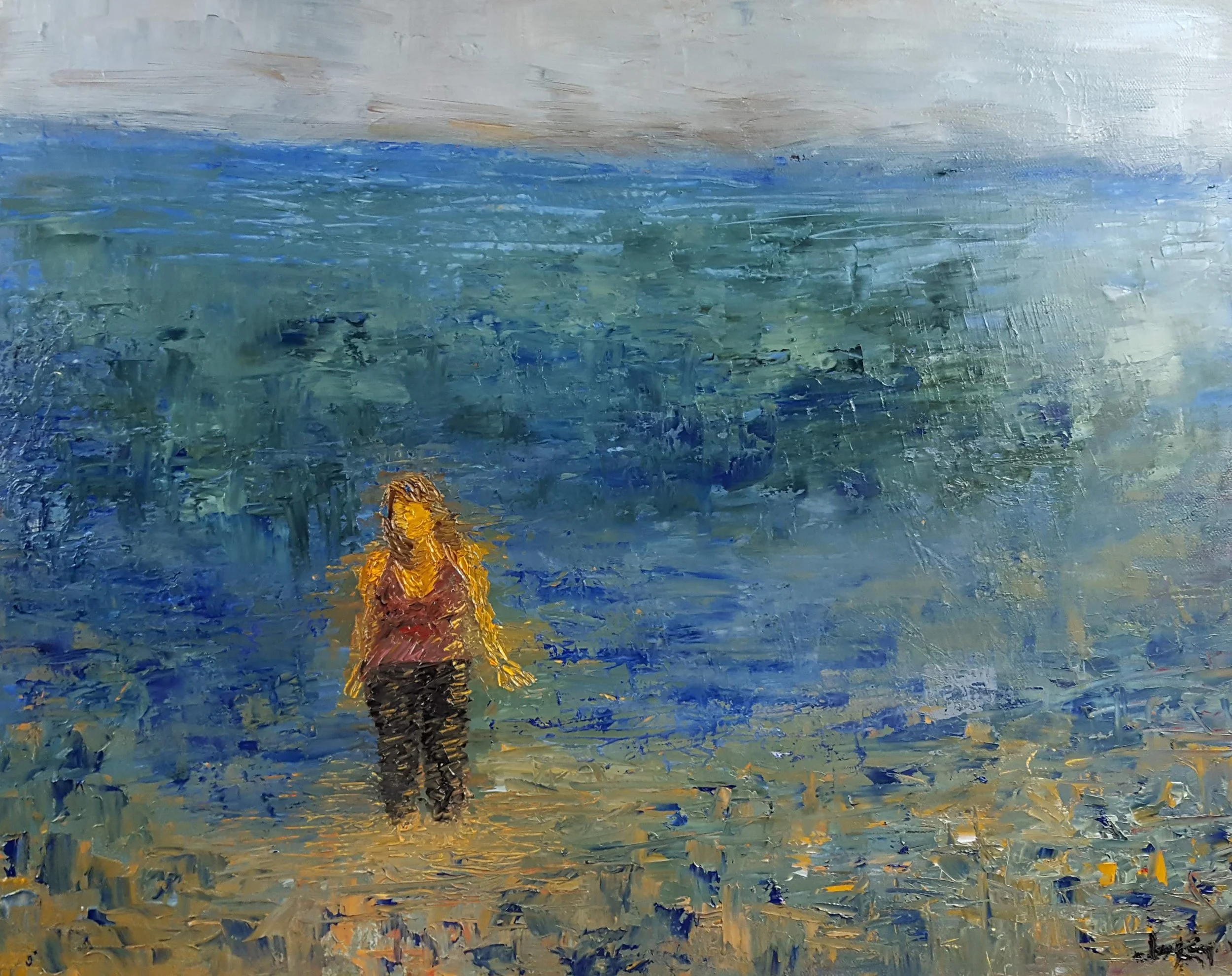 A person with long hair wearing a yellow top and black pants standing on a beach, looking at a large body of water with an overcast sky.