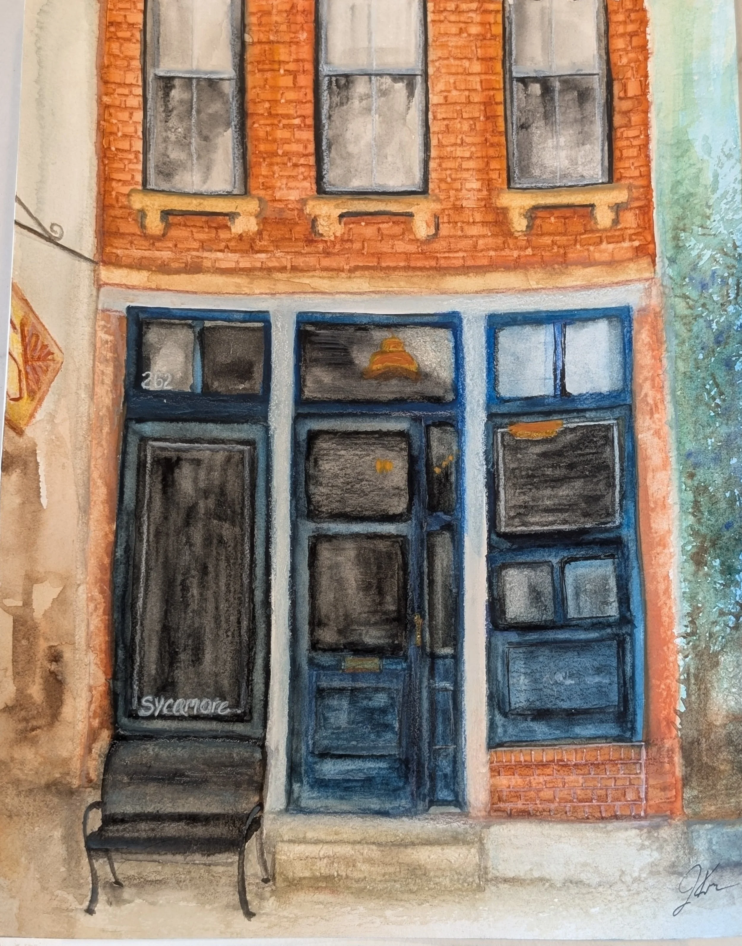 Watercolor painting of a brick building facade with three windows and a door. The door has a sign that reads 'Sylacauga.' There is a bench outside on the sidewalk, and trees are visible on the right side.