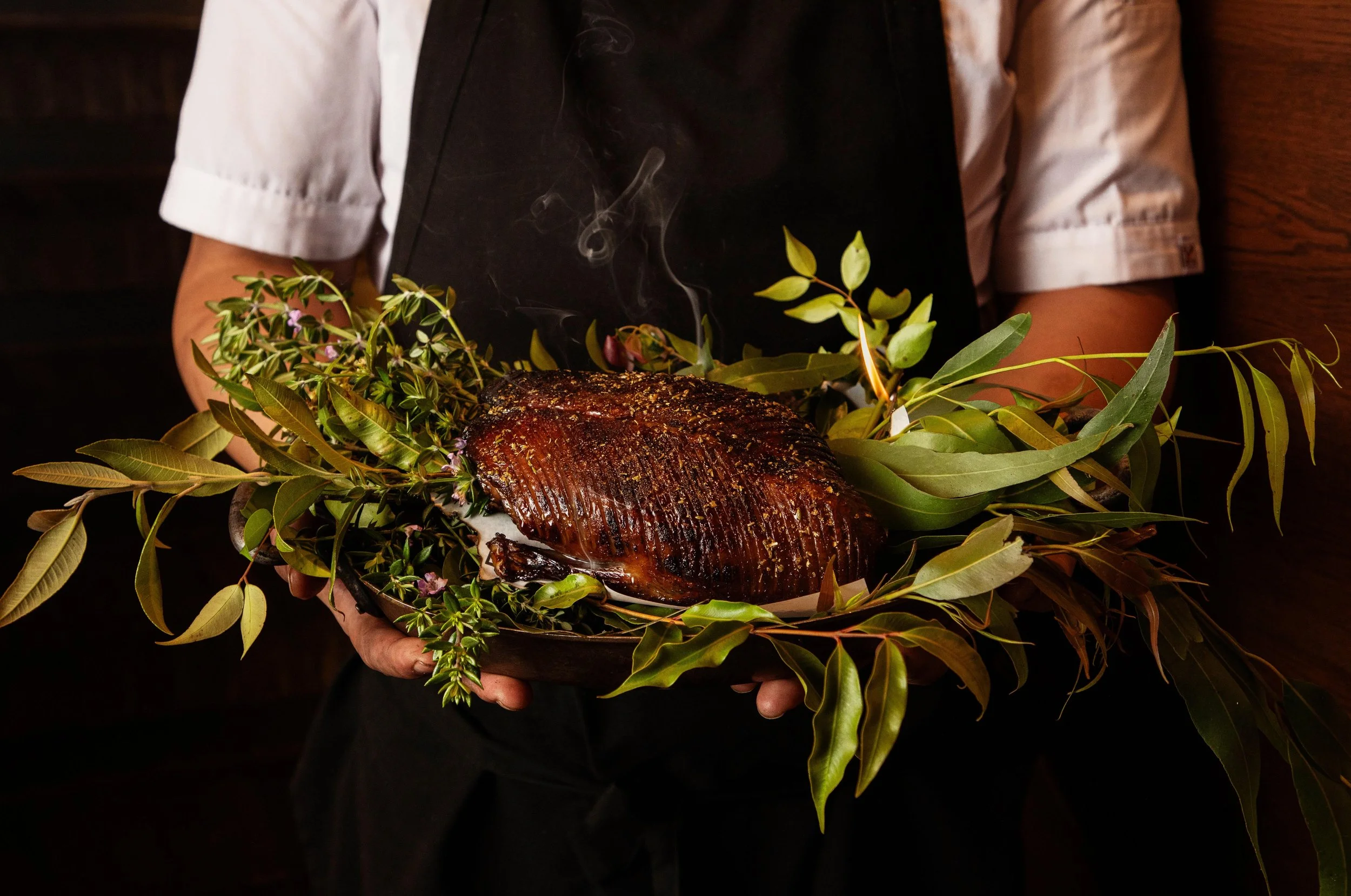 Person holding a roasted piece of meat garnished with green leaves and herbs, with steam rising from it.