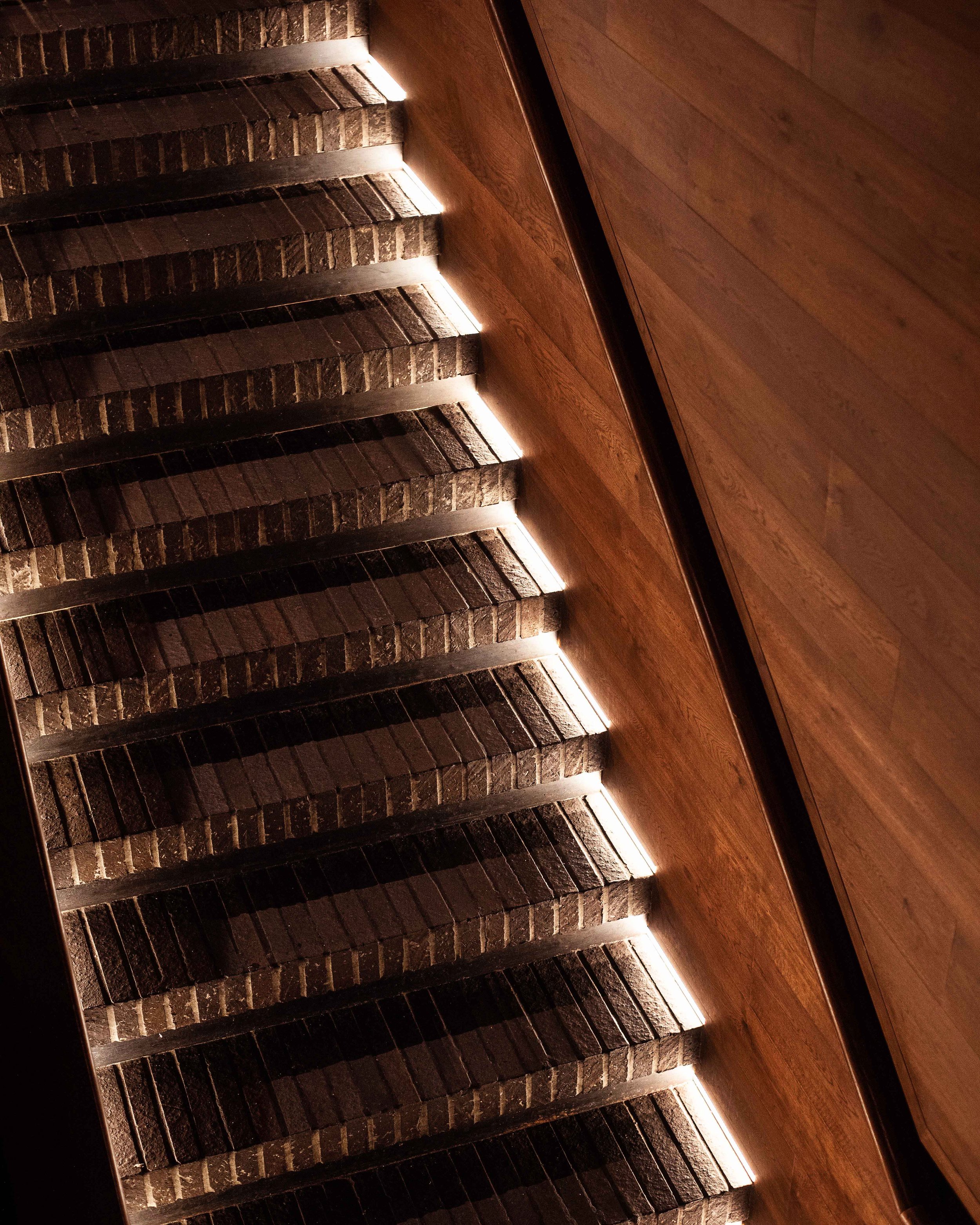 Close-up of a staircase with dark brick treads, illuminated by led lights underneath each step, and a wooden wall on the right side.