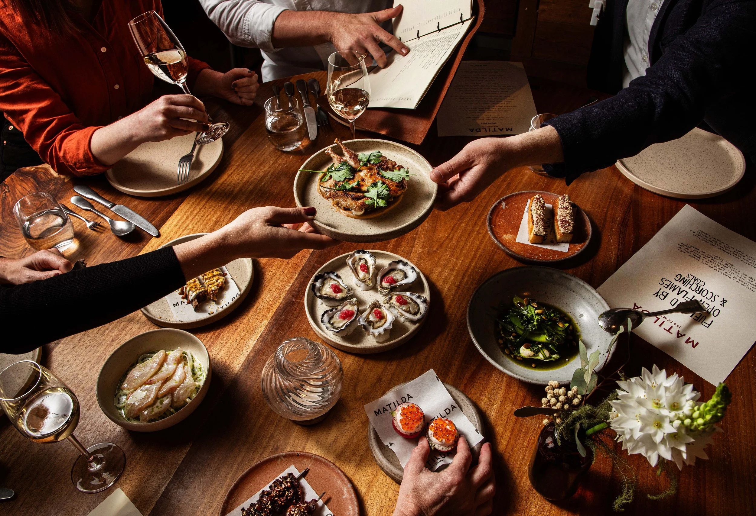 People enjoying a dinner at a wooden table with various dishes, including oysters, mains,, and desserts, with wine glasses and menus.