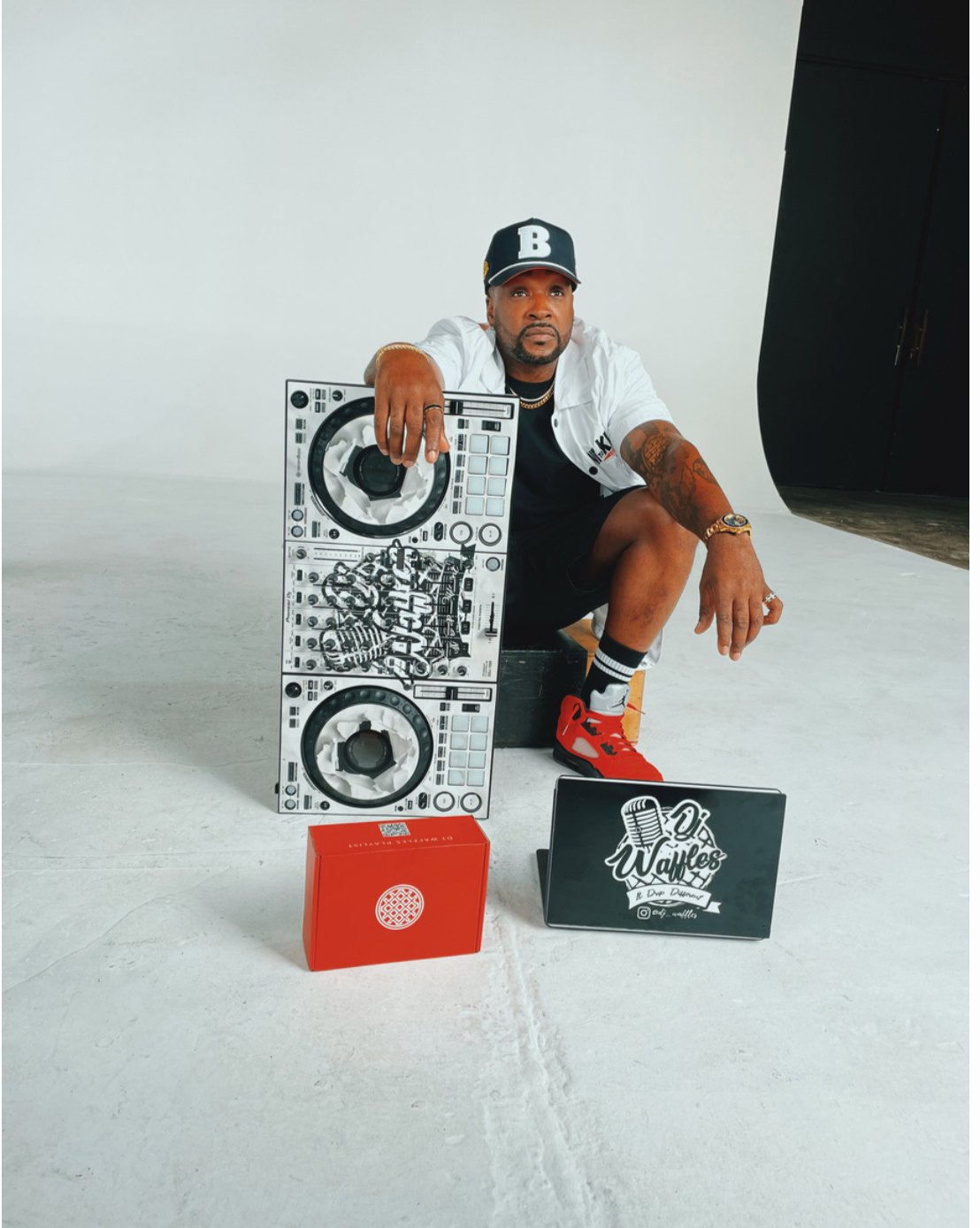 A man crouches in a studio with a large DJ controller and promotional items, including a sign that says "O’ Waffles." He is wearing a baseball cap, white jacket, black shorts, and red sneakers, with tattoos visible on his arms.