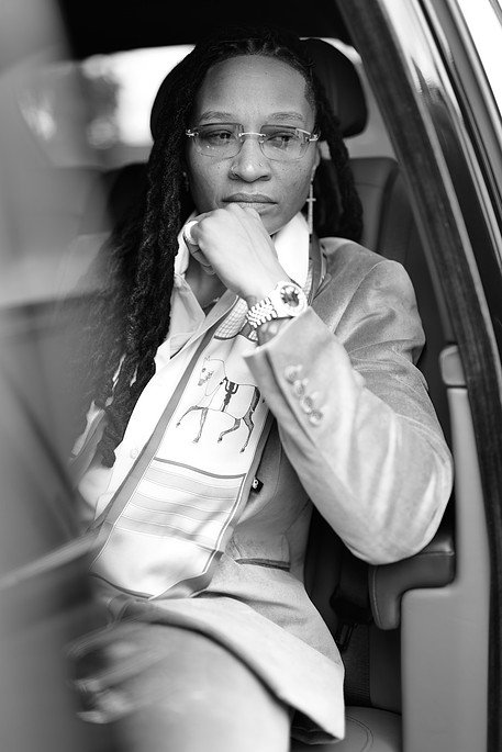 A woman with glasses and dreadlocks sitting in a car, resting her chin on her hand, wearing a jacket and a patterned shirt.