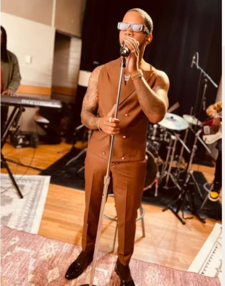 A person wearing sunglasses, a sleeveless brown vest, and brown pants standing in a music studio or rehearsal space, holding a microphone and singing, with a drum set and keyboard in the background.