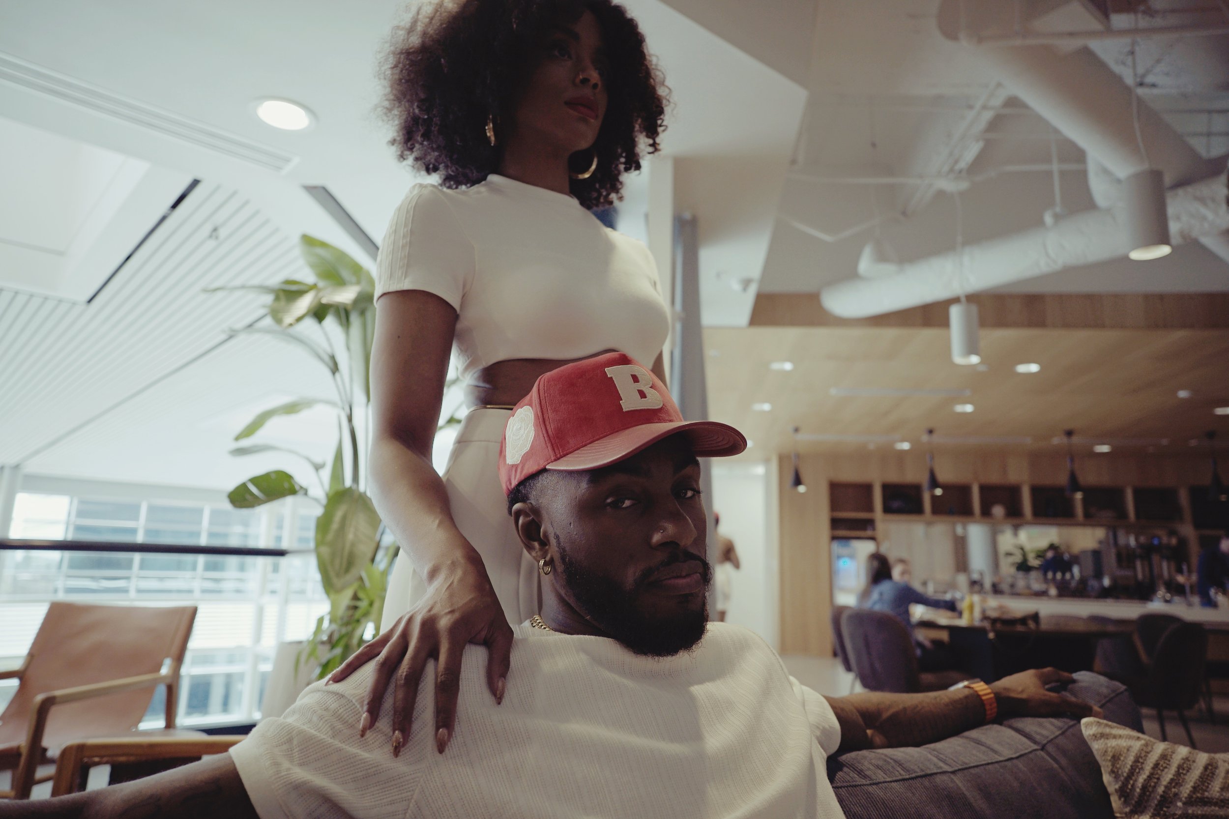 A woman with curly hair and earrings stands behind a man with a beard, wearing a red cap with a letter B and a white shirt with sleeves rolled up in a modern indoor space with large windows, plants, and wooden accents.