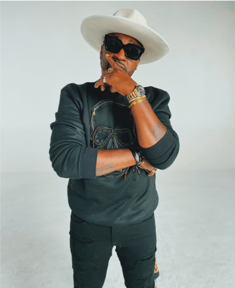 Man wearing large white hat, black sunglasses, black sweatshirt, gold watch, and jewelry, standing with one hand on chin against a plain background.