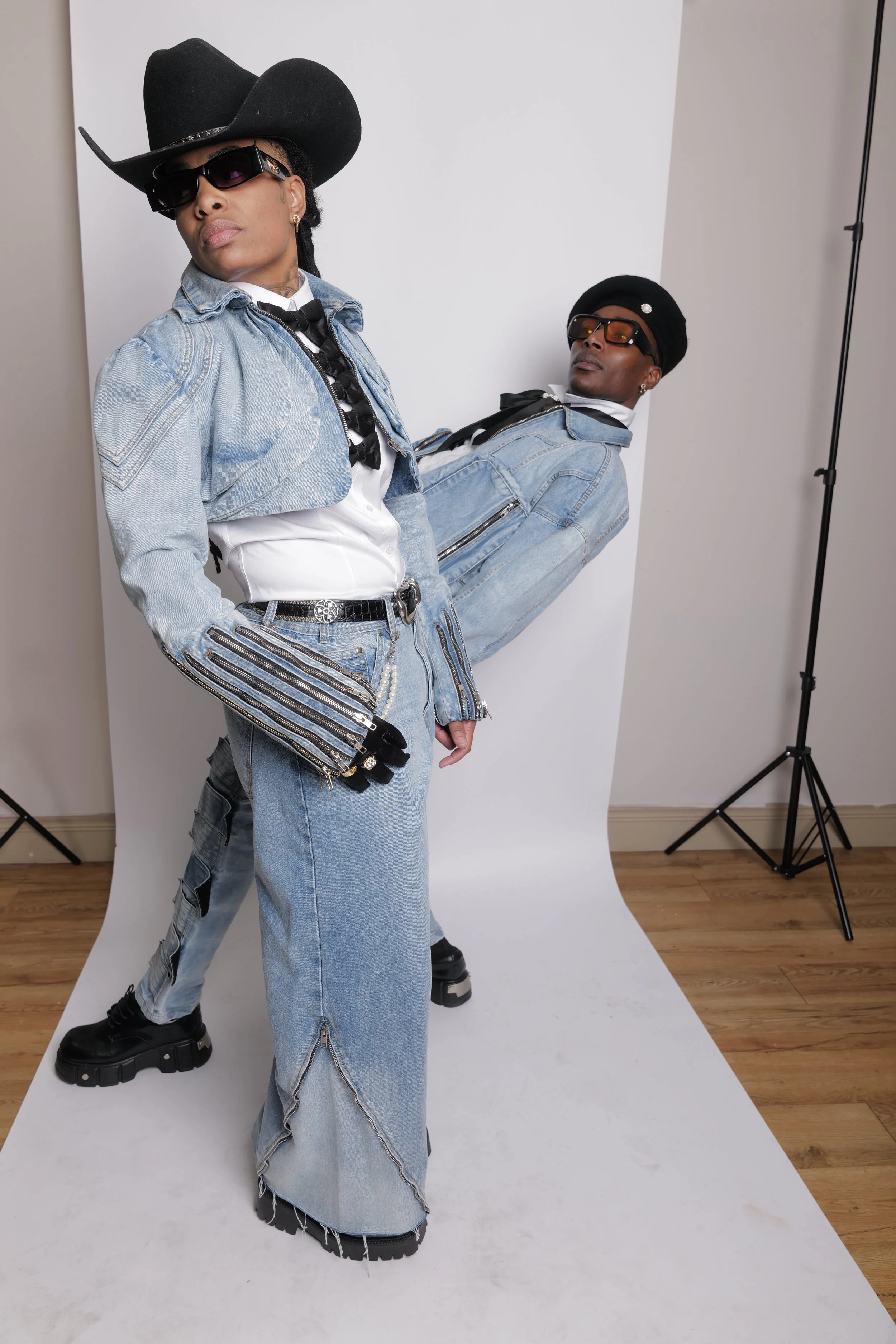 Two people pose in a photography studio against a white backdrop, dressed in denim outfits with accessories. One stands in front wearing a large black cowboy hat, sunglasses, a denim jacket, and wide-leg jeans, while the other leans in the background