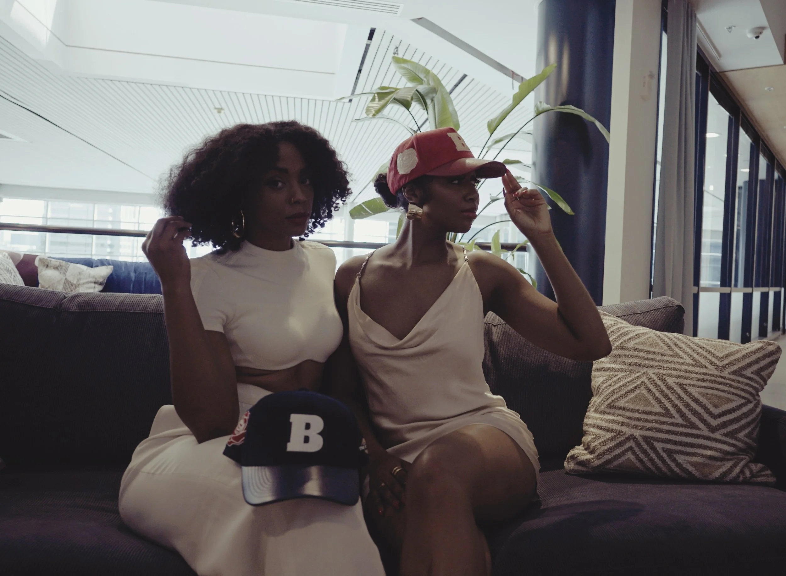 Two women sitting on a dark sofa in a modern indoor setting, with large windows and plants in the background. One woman with curly hair is wearing a white top and pants, with a baseball cap with a red and black design, placed on the sofa in front of her. The other woman, wearing a satin dress and a red baseball cap, is adjusting her cap. There are patterned pillows on the sofa.