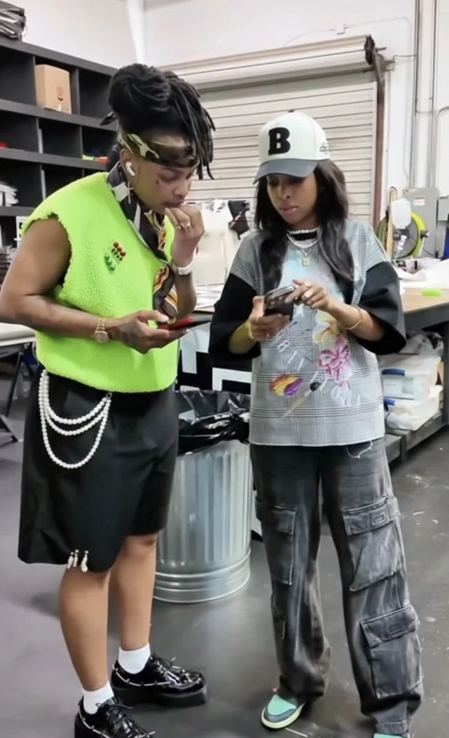 Two women stand in a workshop or storage room, looking at their phones. The woman on the left wears a neon green vest, black shorts with pearl chains, and chunky black shoes. The woman on the right wears a gray cap, oversized gray shirt with a pink and purple design, and baggy cargo pants.