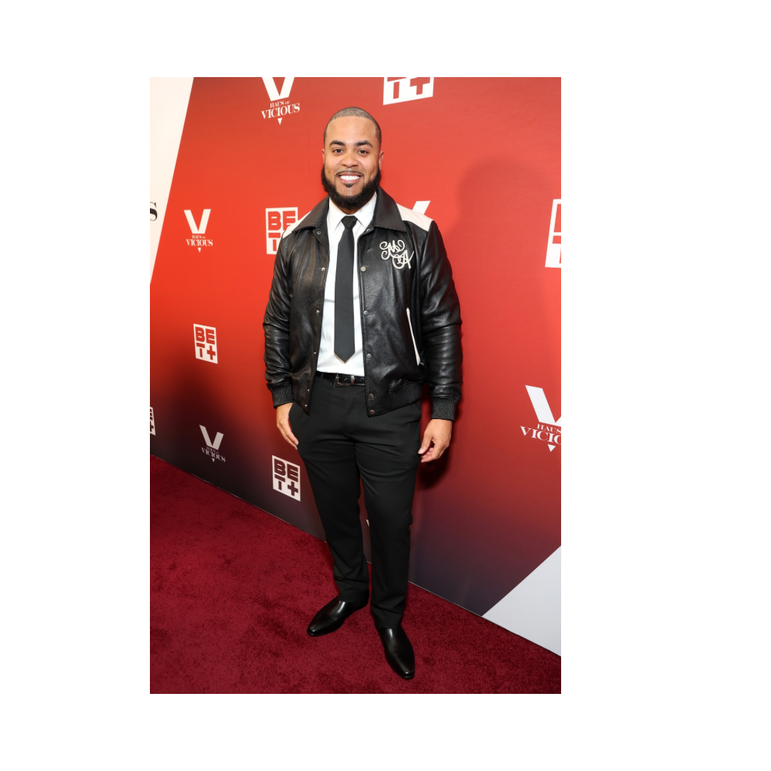 A man with a beard and shaved head smiling, dressed in a black leather jacket, white shirt, black tie, black pants, and black shoes, standing on a red carpet with a red background featuring logos and branding for BET and VICE.