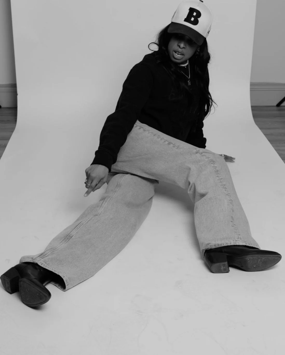 A woman sitting on the floor in a photography studio with a white backdrop, wearing a baseball cap, a dark hoodie, baggy jeans, and boots, posing with a casual expression.