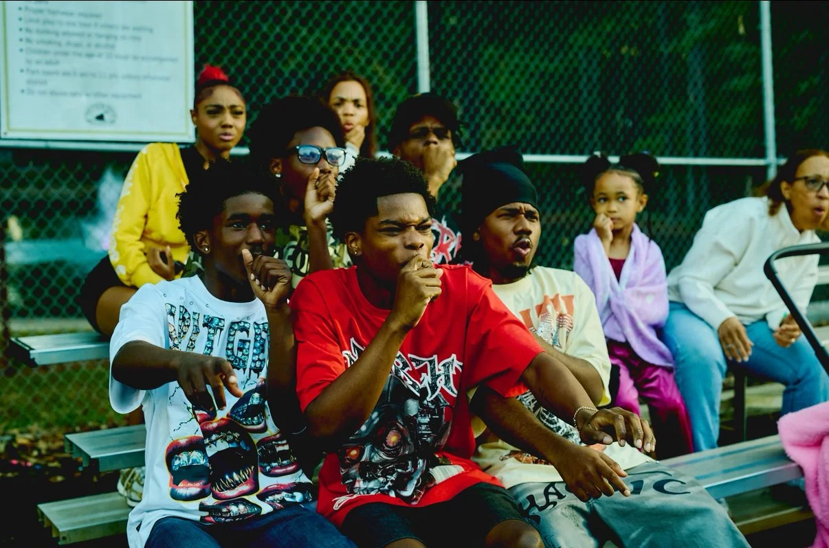Group of young people sitting on bleachers outdoors, reacting with concern or confusion, against a chain-link fence background.