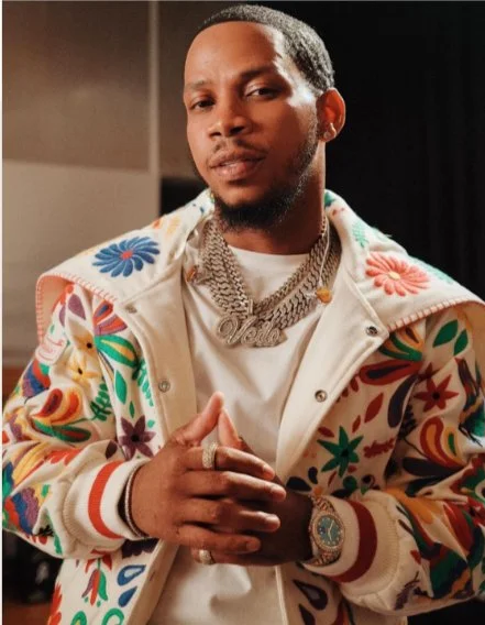 A man with a confident expression wearing a colorful floral jacket, gold chains, and a watch, with his hands clasped together.