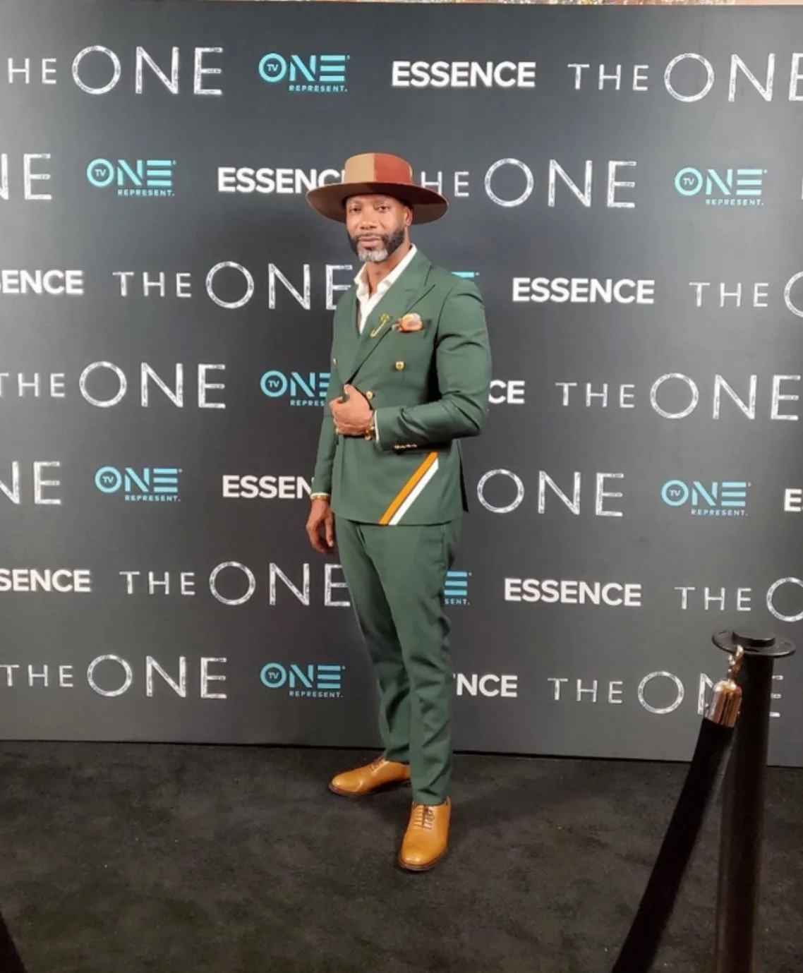 A man dressed in a green suit with gold accents, tan boots, and a wide-brimmed hat, standing on a black carpet in front of a backdrop with logos and text including "THE ONE," "ESSENCE," and "TV ONE."