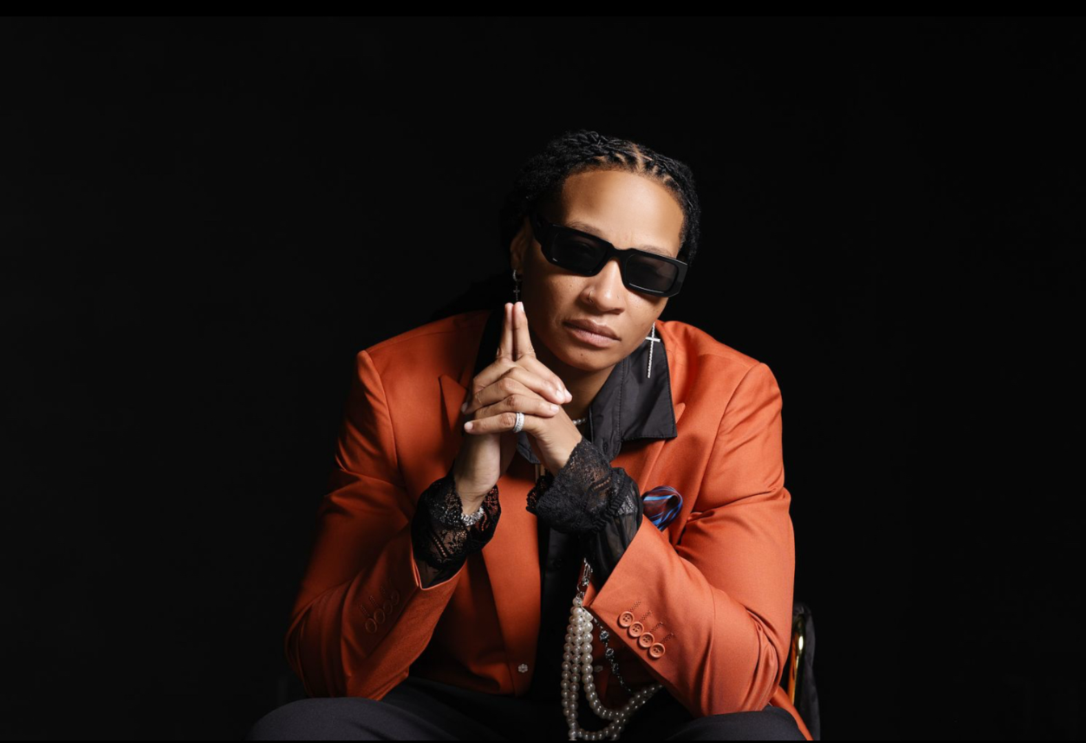 A woman wearing black sunglasses, an orange blazer, black lace gloves, and jewelry, posed against a black background.