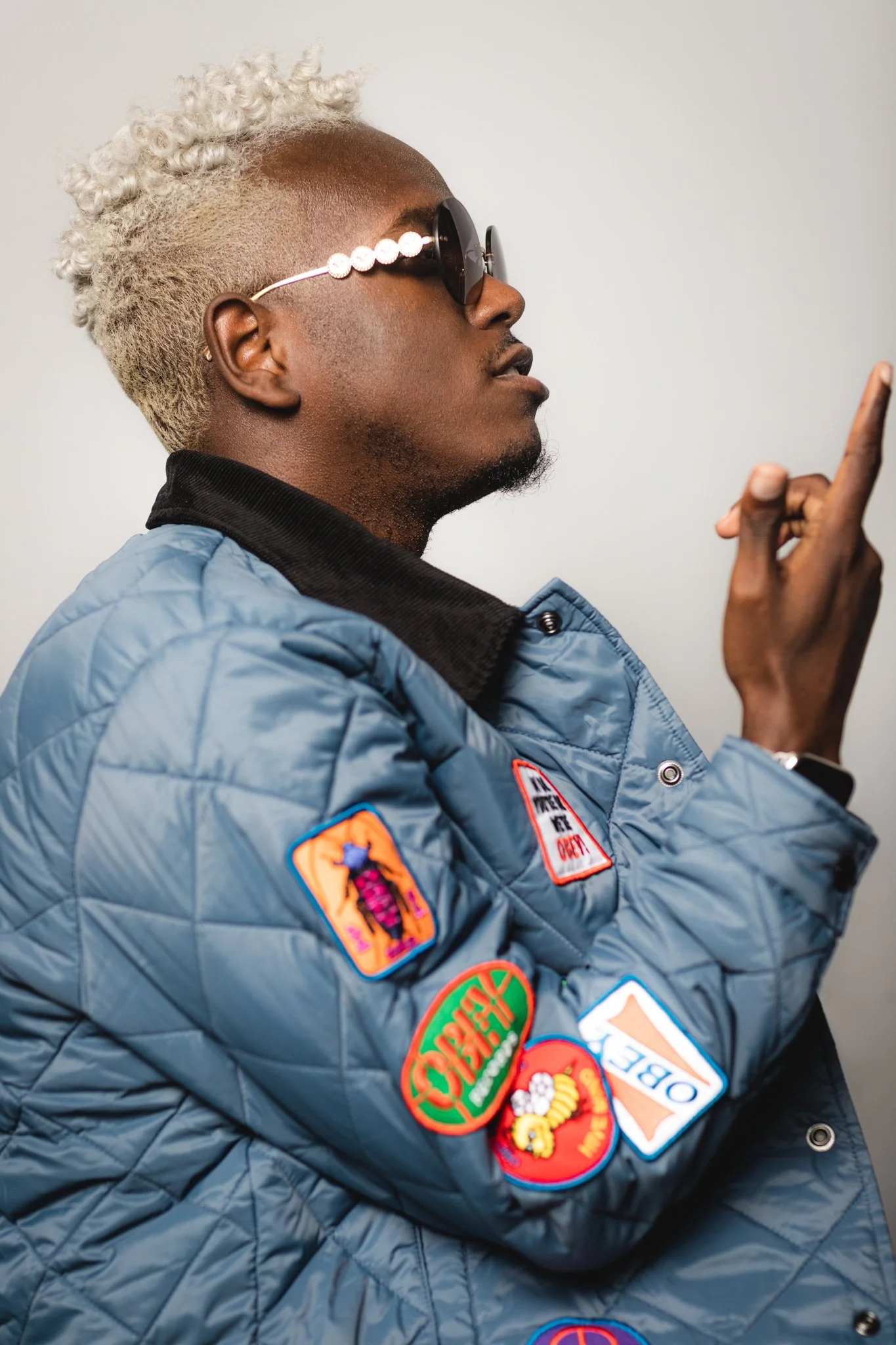 A young man with light blonde curly hair and sunglasses features a blue quilted jacket with various colorful patches, and he is pointing with his finger.