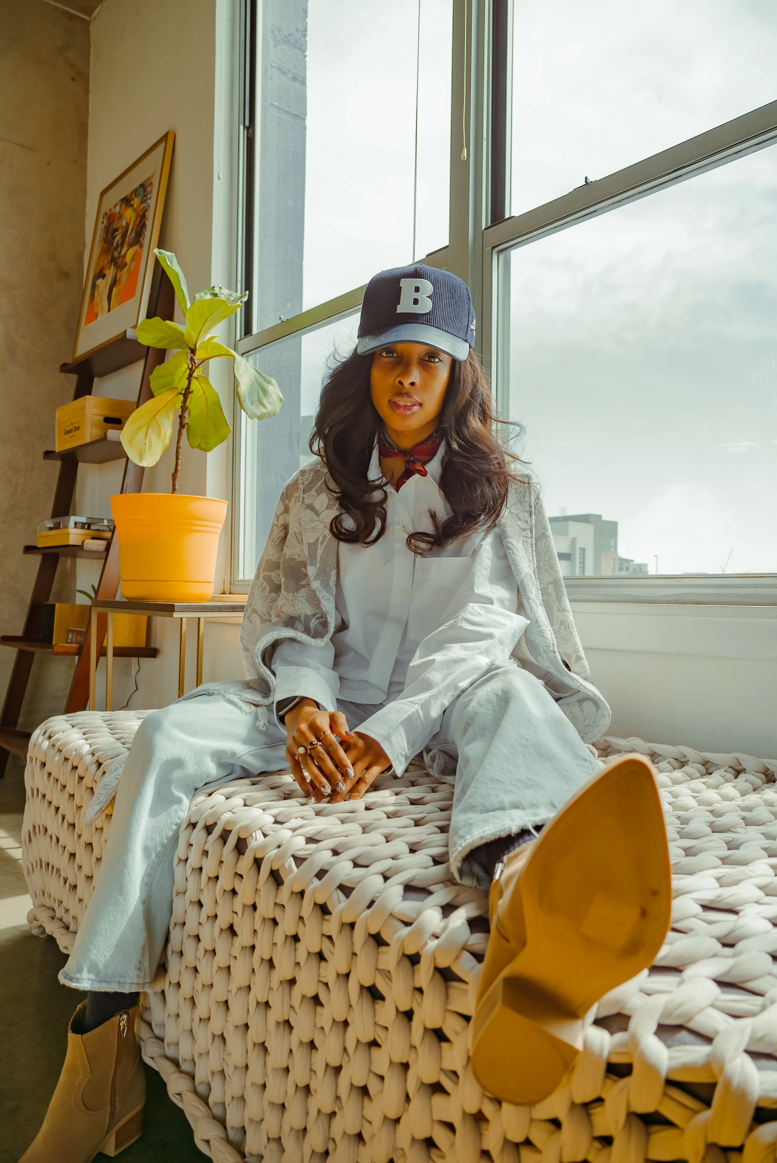 Woman sitting on a woven bench near large windows, wearing a black baseball cap with a white 'B', a white shirt, light-colored pants, and tan boots, with a scarf around her neck, in a modern indoor space with a plant and artwork
