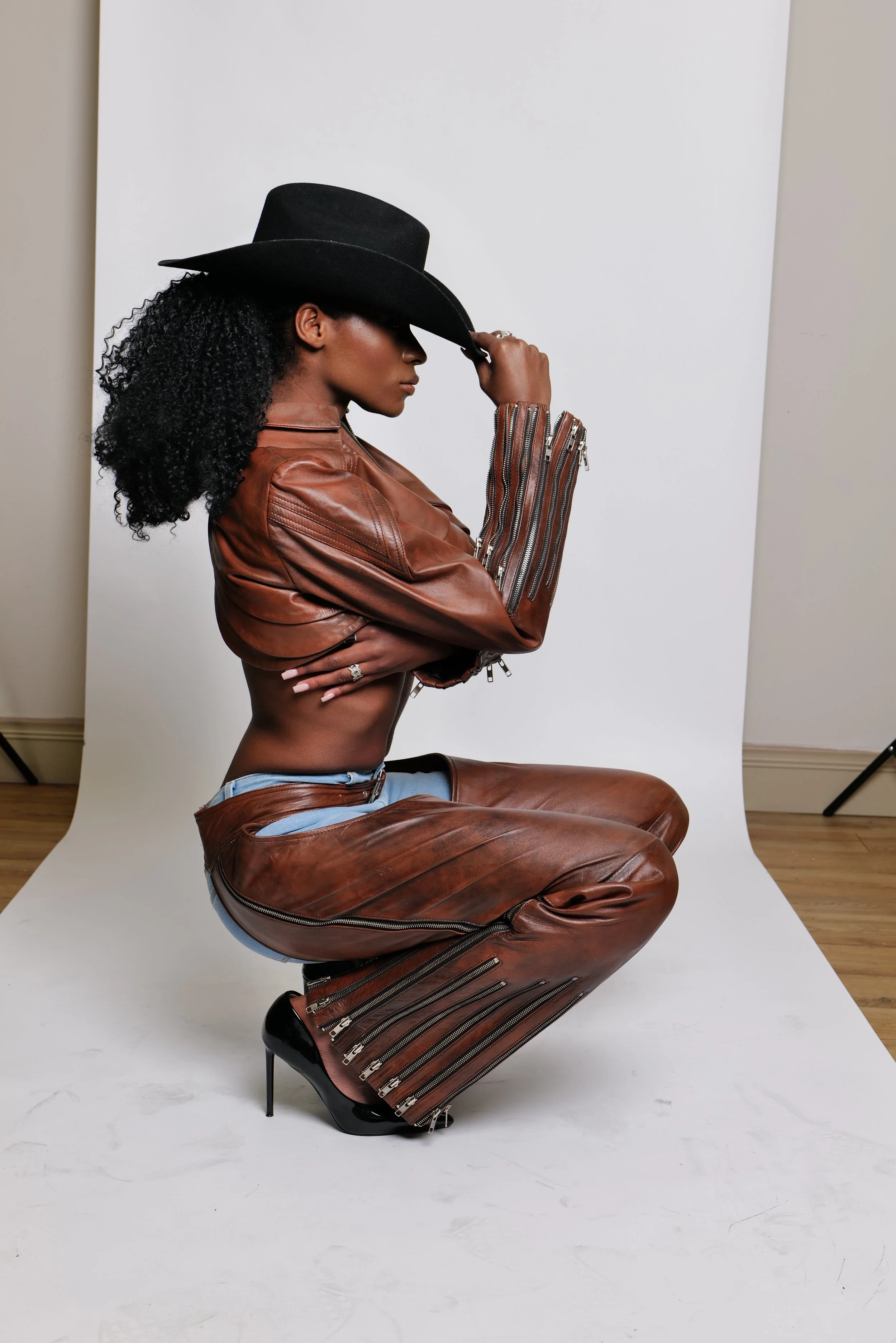 A woman wearing a black wide-brim hat, brown leather jacket, and brown leather pants with zipper details, is kneeling on a white backdrop and posing with her hand adjusting her hat.