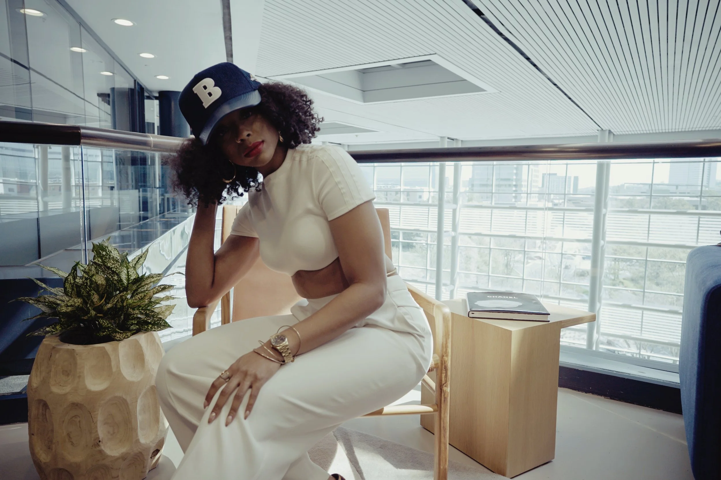 A woman with curly hair wearing a baseball cap, cream-colored crop top, and white pants, sitting on a wooden chair in a modern building with large windows, a potted plant, and a small wooden table with books.