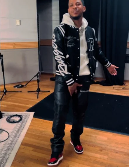 A man standing in a recording studio, wearing a black and white varsity jacket, black leather pants, and red and white sneakers, with microphones and recording equipment visible in the background.