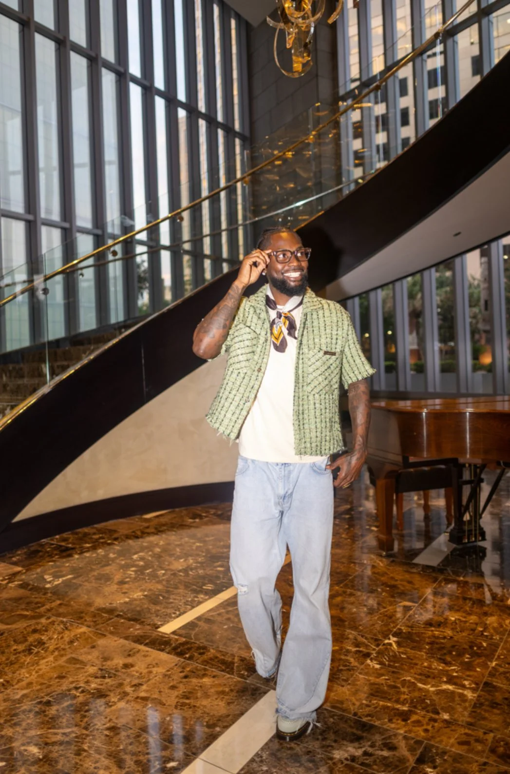 A man with glasses and a beard smiling and adjusting his glasses inside a modern building with large glass windows, a black and gold staircase, and a piano in the background.