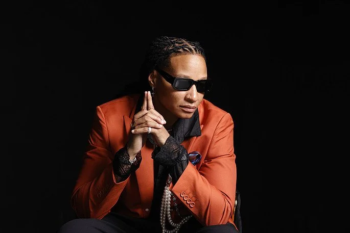 A woman with braids wearing sunglasses, an orange blazer, black lace gloves, and pearl jewelry, posed against a black background.