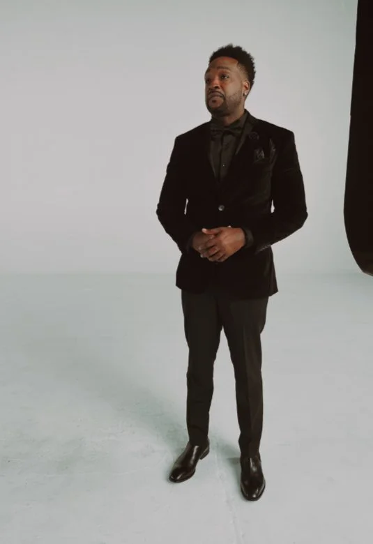 A man dressed in a black tuxedo with a black shirt and shiny dress shoes, standing against a plain background.