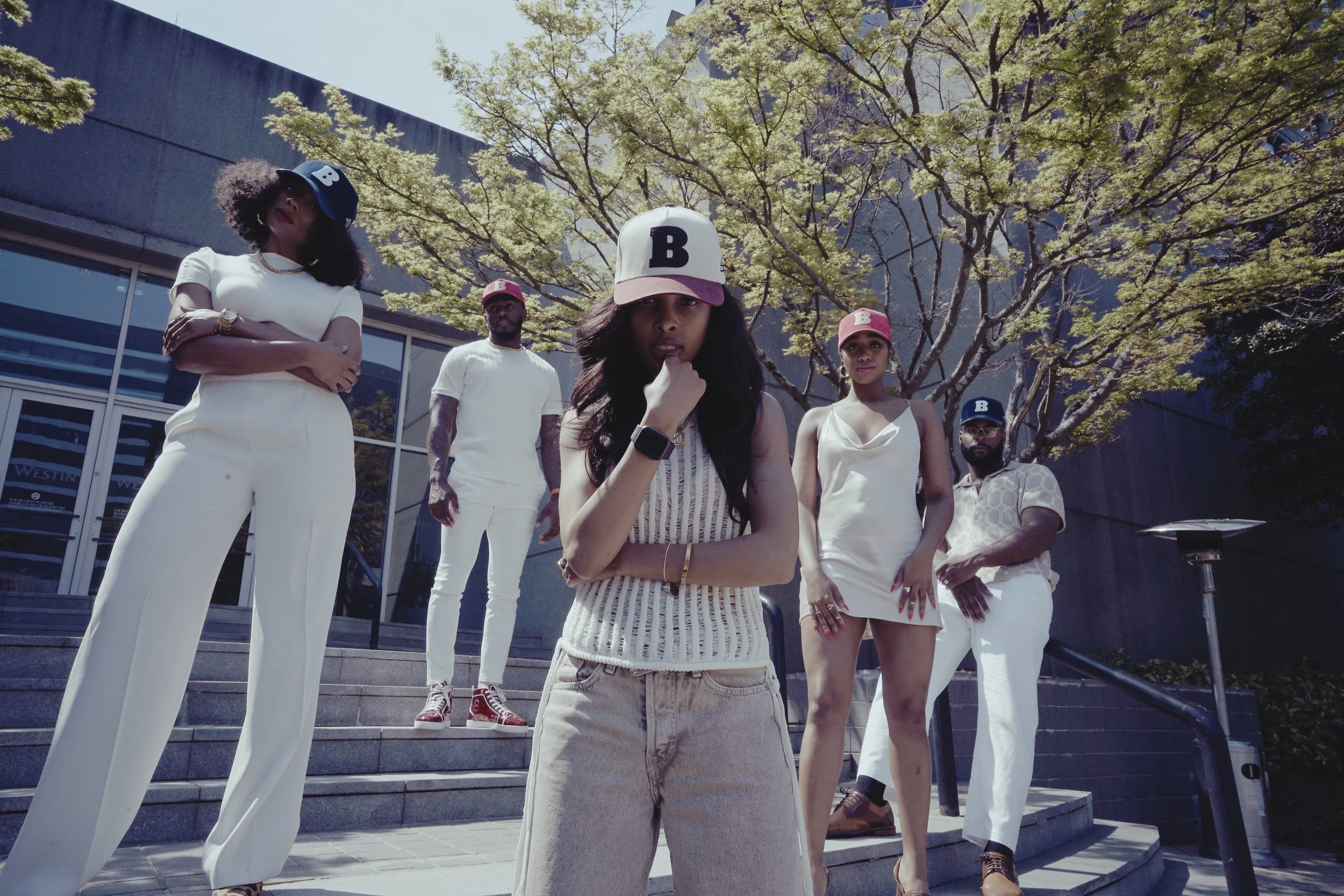 A group of five fashionable people standing on steps outside of a modern building with glass windows and a tree in the background. The women are wearing stylish white and cream outfits, some with baseball caps, and the men are dressed casually, some 