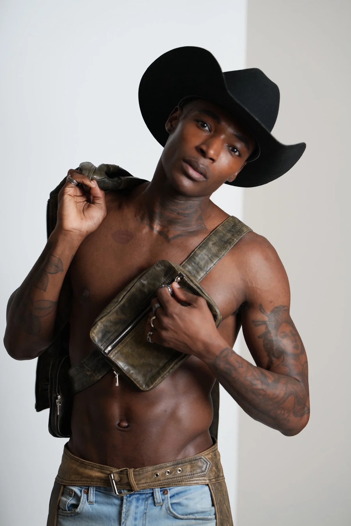 A shirtless Black man wearing a large black cowboy hat, carrying a brown leather bag over his shoulder, and wearing jeans, standing against a plain background.