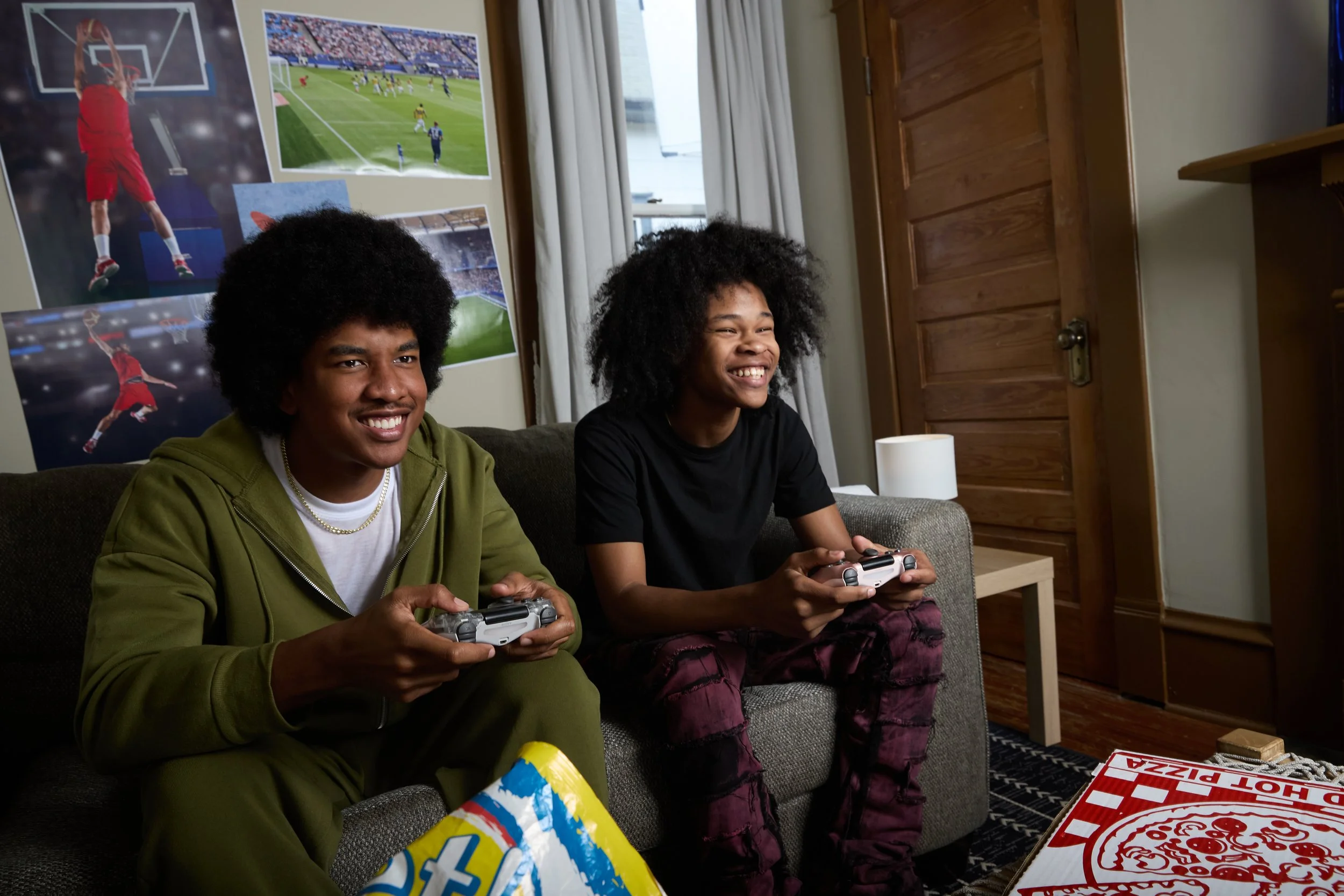 Two young men with curly hair sitting on a gray couch playing video games, smiling and holding controllers, in a room decorated with sports posters, a wooden door, and a pizza box on the table