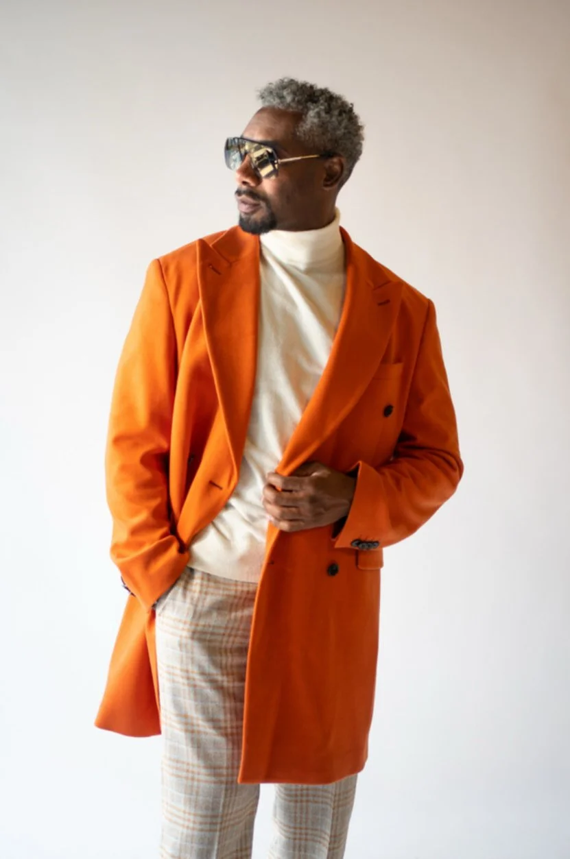 A man with gray hair and sunglasses, wearing a white turtleneck, orange coat, and plaid pants, standing against a plain background.