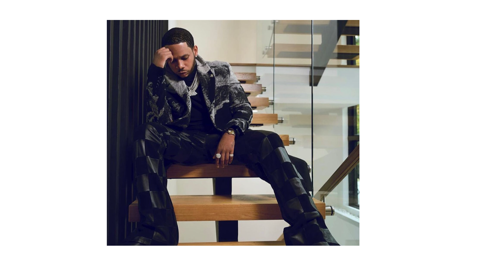 A man sitting on a wooden staircase indoors, looking down with a serious expression, wearing designer clothing and jewelry, with a modern interior background.