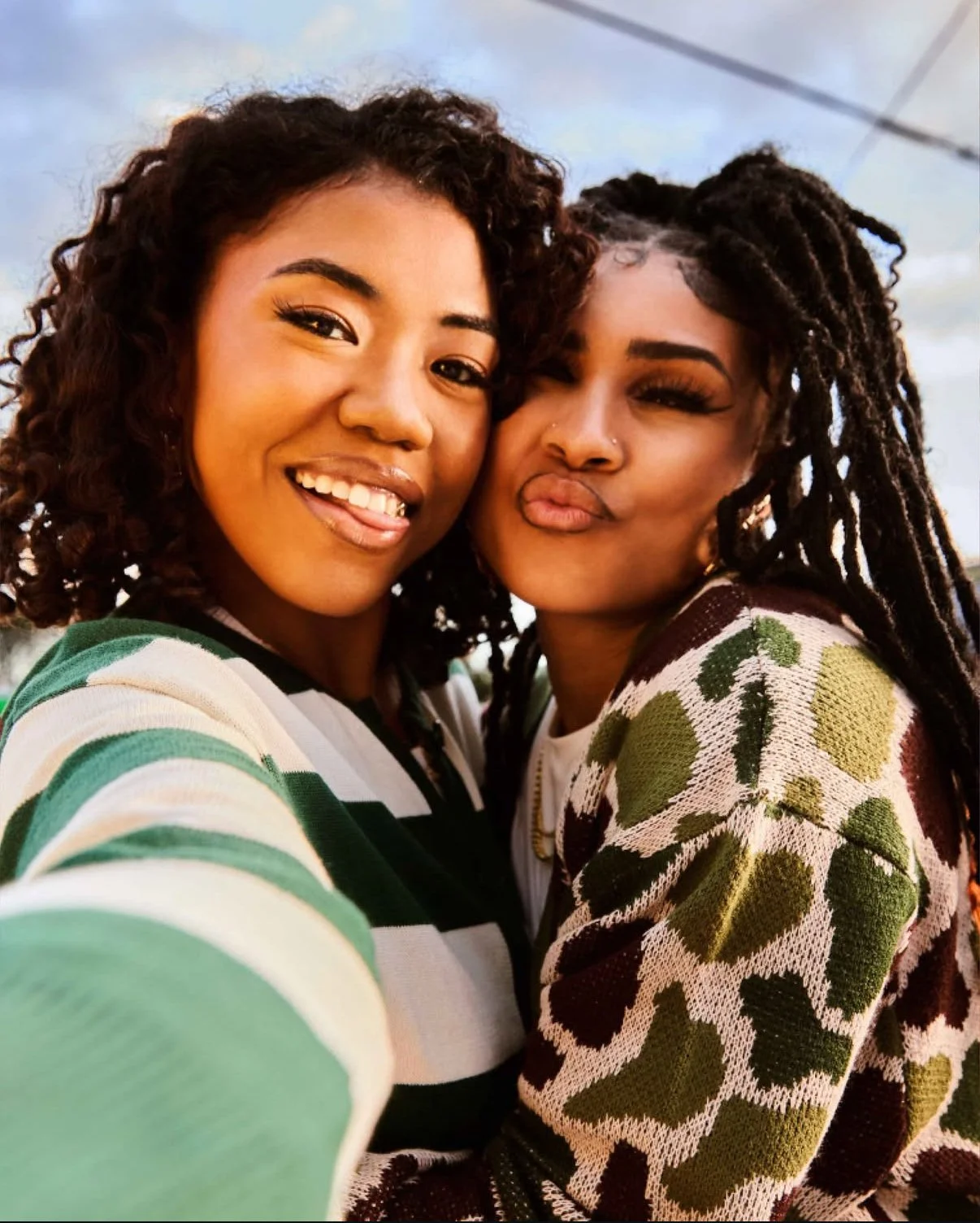 Two women taking a close-up selfie outdoors, smiling and making playful expressions, with a cloudy sky in the background.