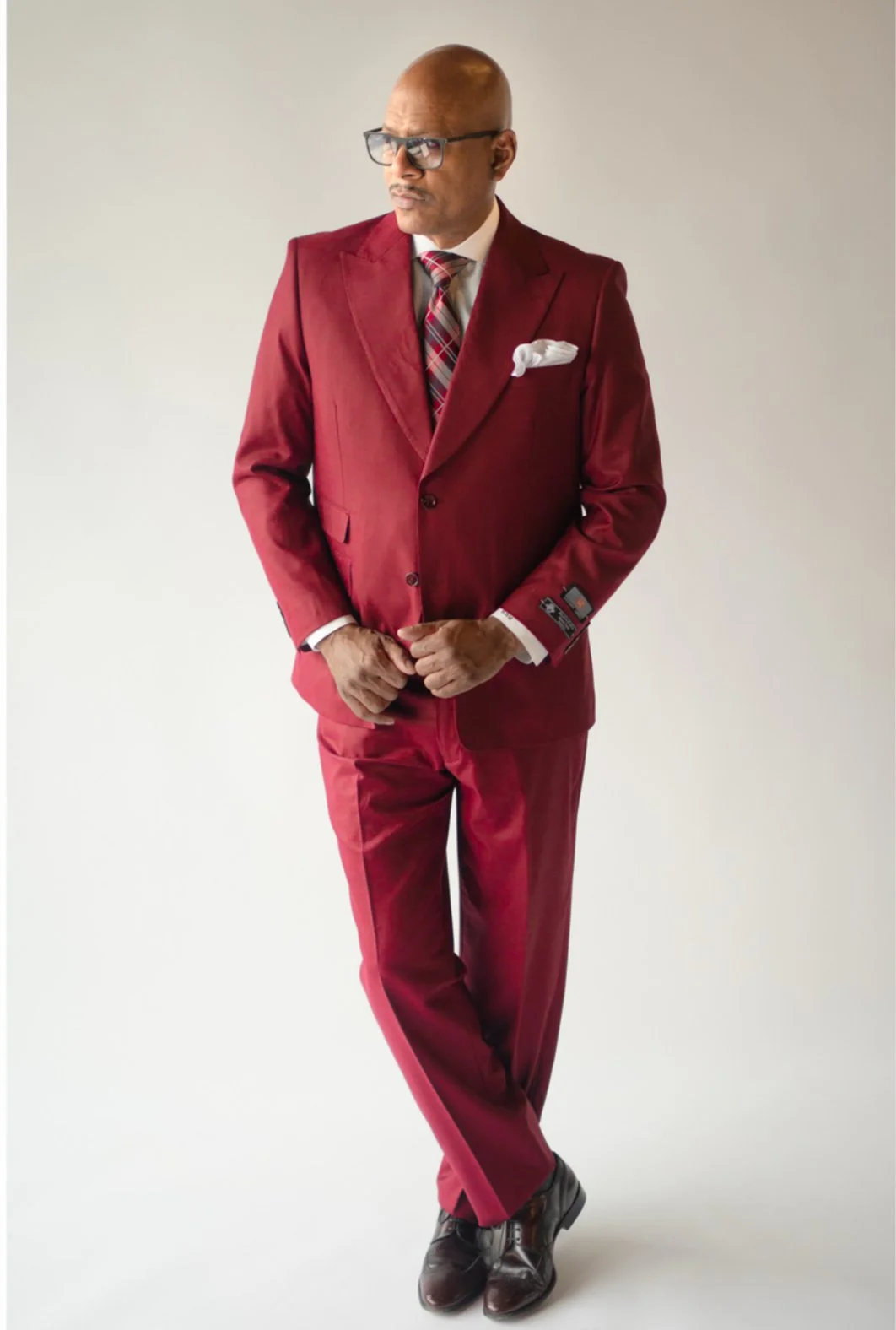 A man dressed in a burgundy suit, with a tie and white pocket square, wearing glasses, standing with crossed legs and hands clasped in front of him against a neutral background.