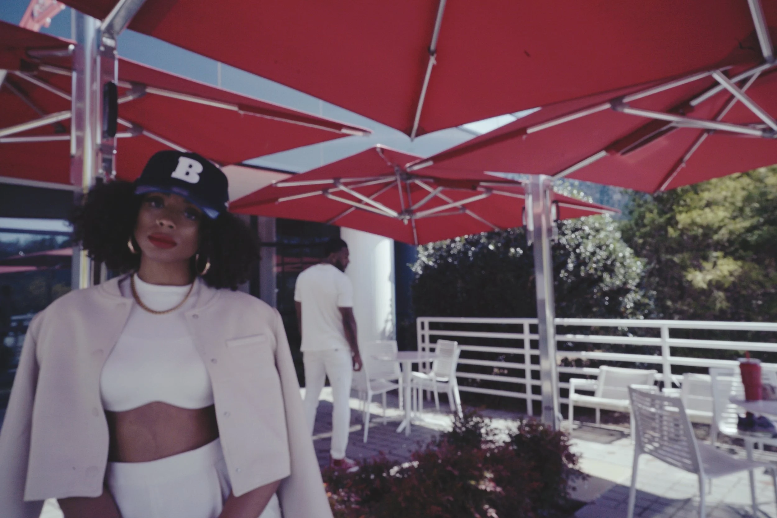 Young woman with curly hair wearing a black baseball cap with a white 'B', white outfit, and red lipstick, standing under red umbrellas at an outdoor dining area with chairs and tables, and a man in white in the background.