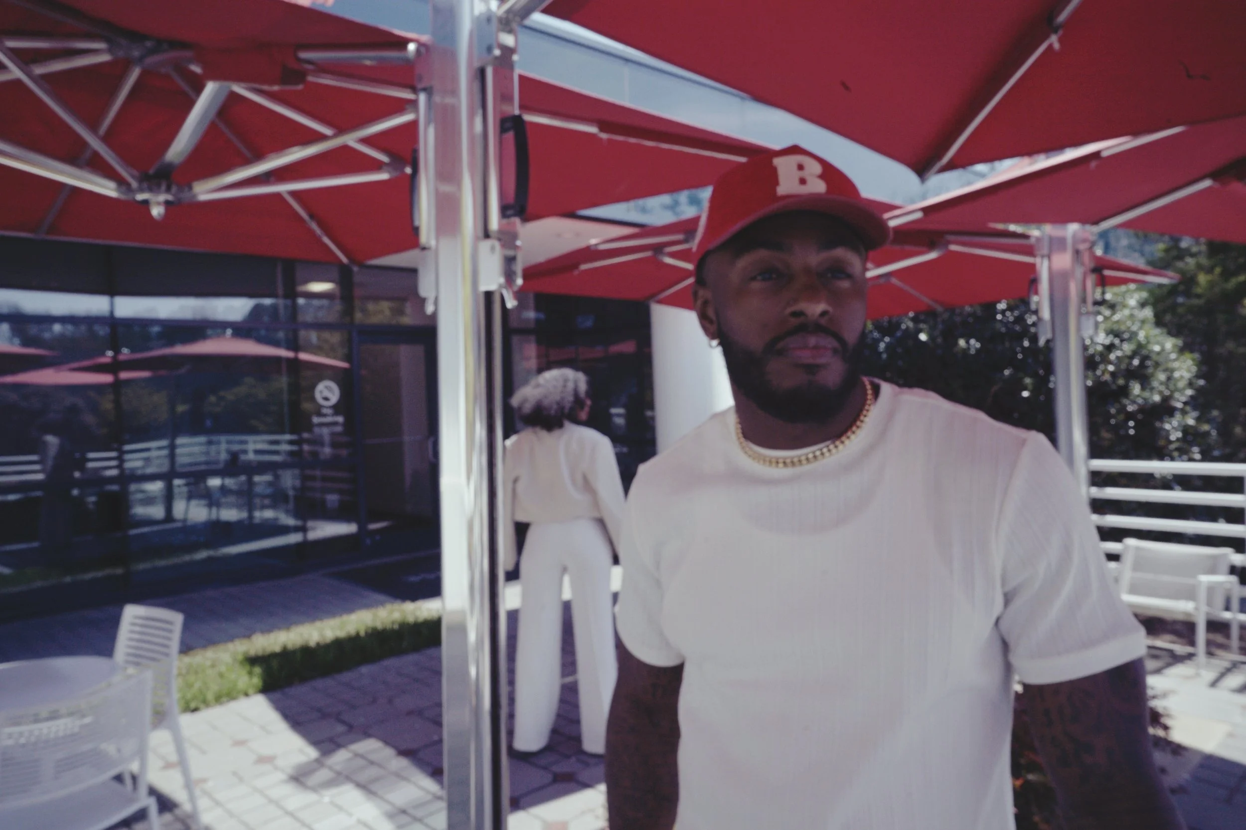 A man wearing a white t-shirt, gold chain, and red baseball cap with a letter 'B' standing under a red patio umbrella outside a building.