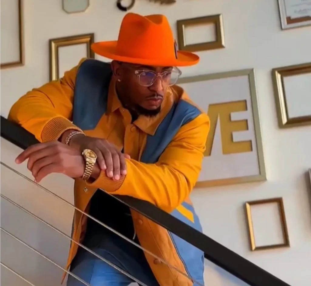 A man wearing glasses, a gold watch, an orange hat, and a colorful jacket leaning over a railing, with a wall in the background decorated with various empty frames and wall art.