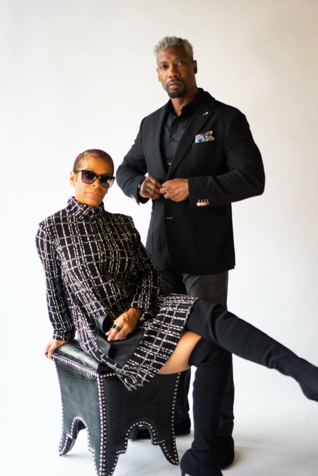 Two stylish individuals posing against a plain white background. The woman is seated on a black stool, wearing sunglasses, a black and white patterned dress, with knee-high black boots. The man stands beside her, wearing a black suit, with a pocket s