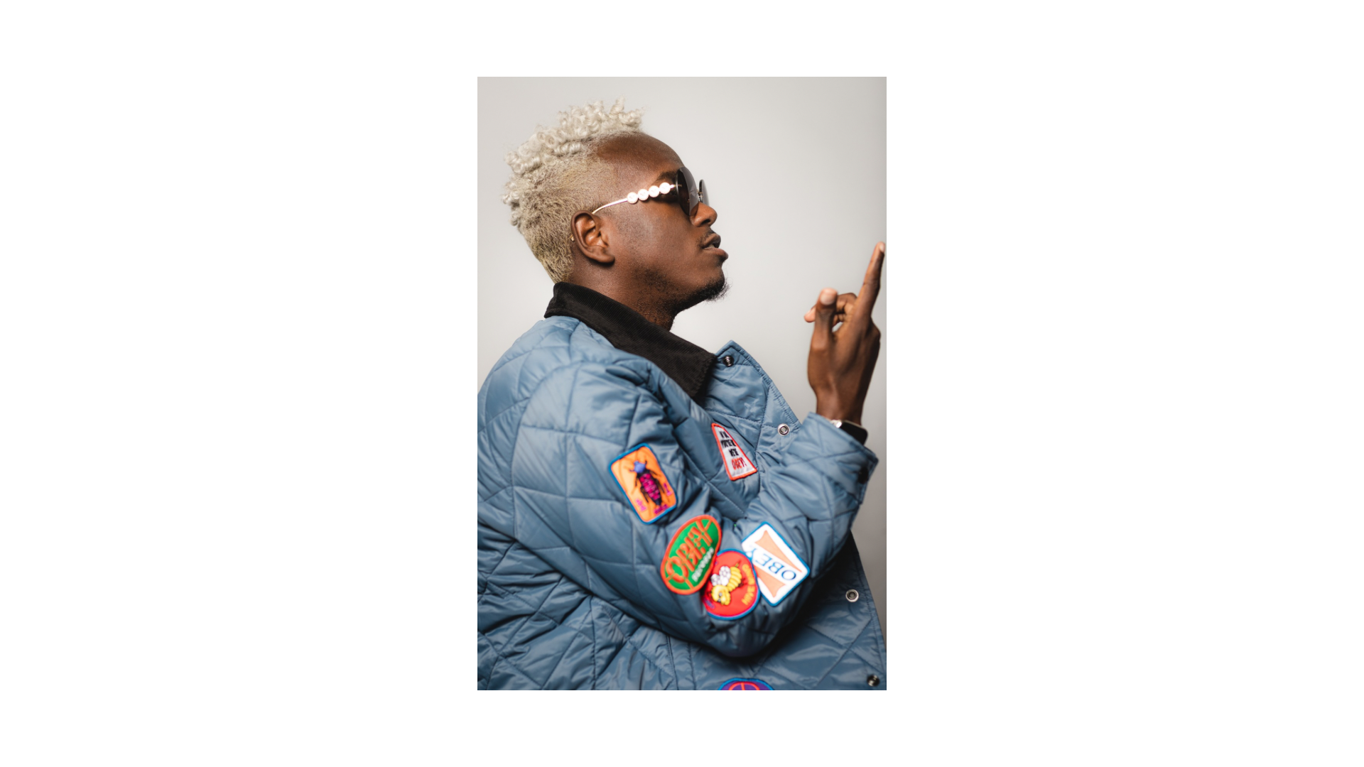A young man with styled blonde curly hair, wearing sunglasses and a blue quilted jacket with colorful patches, poses against a plain background, making a gesture with his right hand.