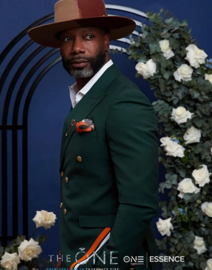 A man wearing a wide-brimmed brown hat, dark green double-breasted blazer, white shirt, and pocket square, standing in front of a dark blue background decorated with white roses.